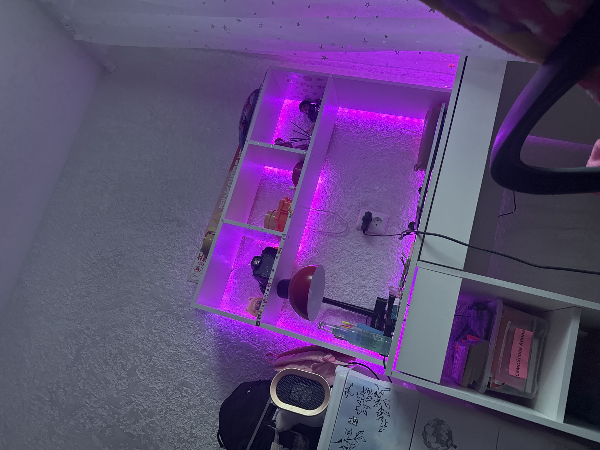 The image shows a desk and shelving unit illuminated by purple LED lights. On the desk, there is a red desk lamp, a bottle of liquid, and other small objects. The shelving unit above the desk is filled with various items, including books, a camera, and decorative objects. To the left of the desk, there is a white dresser with a small portable heater on top. The room has textured walls and white curtains with star patterns. The overall scene depicts a personal workspace or bedroom setting.