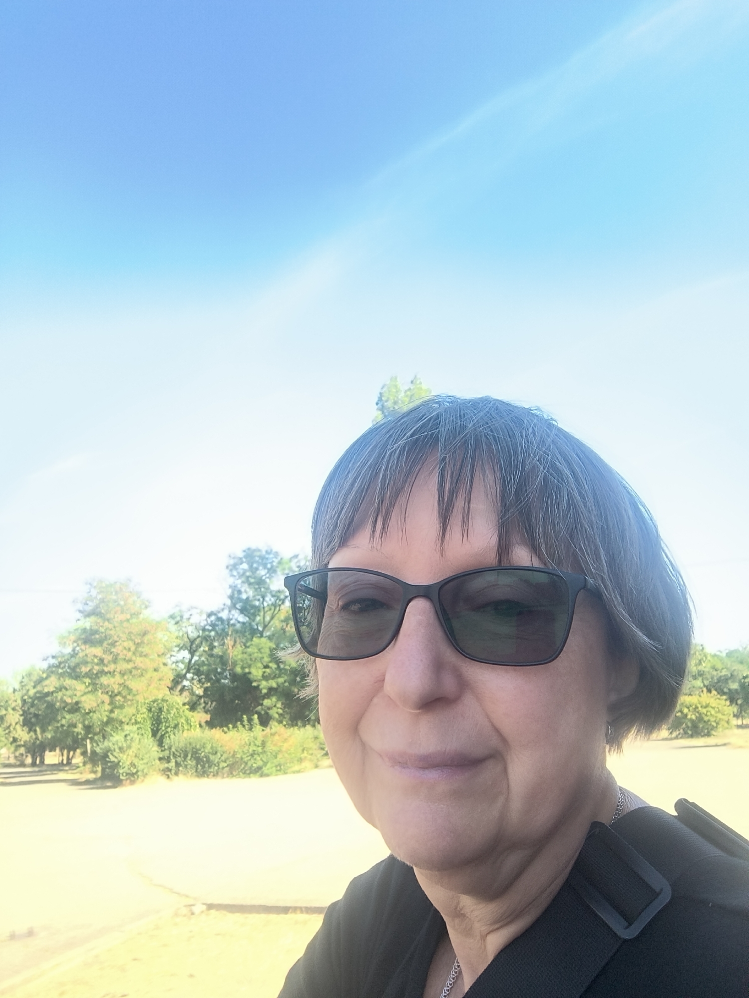 A woman with gray hair and glasses takes a selfie outdoors. She is wearing a black shirt and a silver necklace. Behind her, there are trees and a bright blue sky. The location is not identifiable as Mykolaiv, Ukraine from the image.