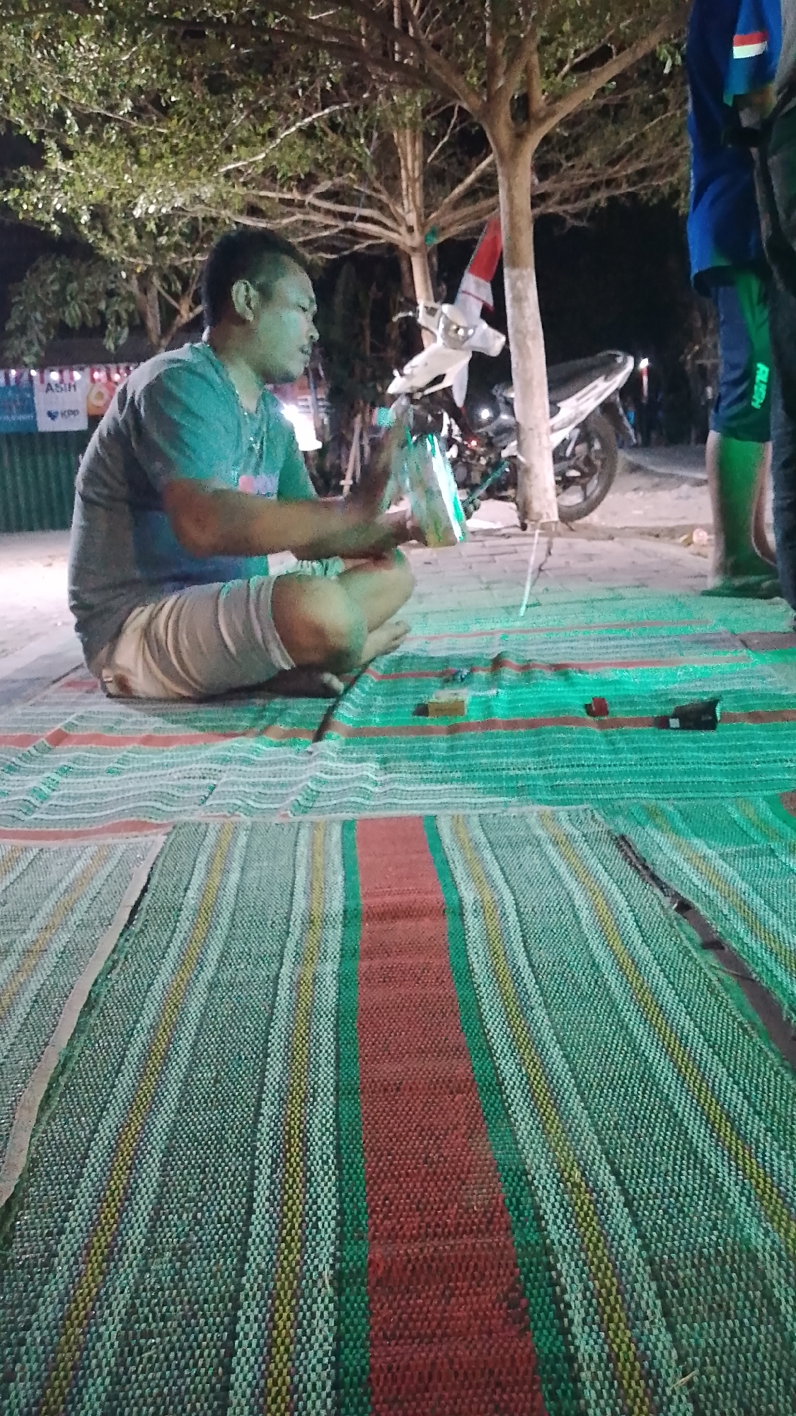 In Karanglangu, Indonesia, a man sits cross-legged on a woven mat in front of a tree. He wears a teal t-shirt and light-colored shorts, with his hands holding a translucent, possibly plastic, object. A white motorcycle with red and white decorations on its handlebars is parked behind him. To the right, another person, seen from the waist down, wears a blue shirt and dark shorts with a green stripe. The environment appears to be outdoors, with trees and foliage visible in the background, suggesting a park or roadside setting illuminated by artificial light, indicating it is nighttime. The ground is covered with a large, striped woven mat.