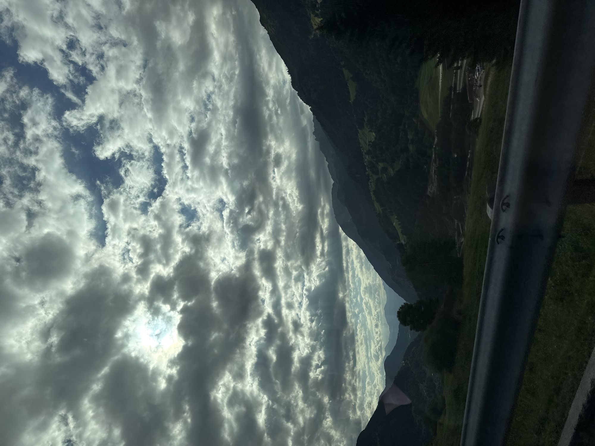 The image captures a dramatic mountainous landscape under a cloudy sky, with the sun partially visible through the clouds, casting rays of light. The foreground features a dark metal guardrail, likely on a road or scenic overlook, with green grass below. Beyond the guardrail, rolling hills covered in dense green trees ascend into steep, dark mountains. In the distance, layers of mountains fade into a hazy blue. The sky is filled with heavy, dark clouds mixed with patches of brighter, lighter clouds, creating a dynamic and somewhat dramatic atmosphere. The scene suggests a location in a mountainous region, likely Airolo, Switzerland, known for its alpine scenery. There are no people or specific objects other than the guardrail and natural elements visible.