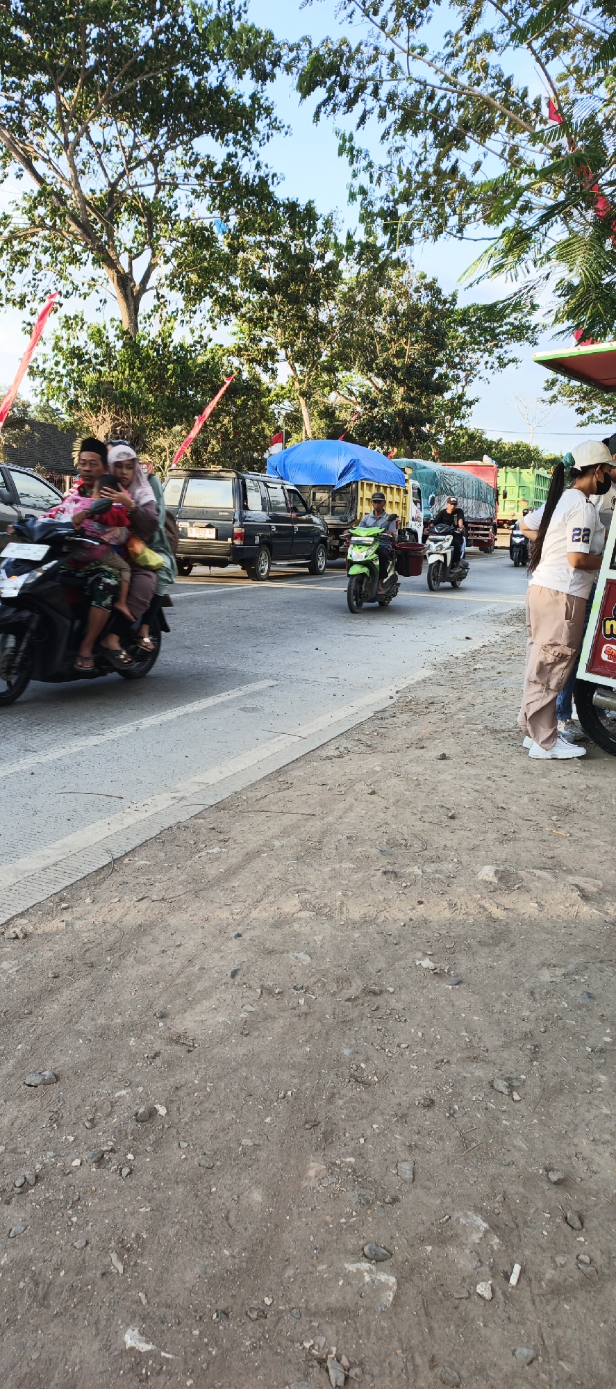In Kasiyan Timur, Indonesia, this image captures a traffic scene with several vehicles on a paved road. A motorcycle in the foreground carries a family of four: a driver, two passengers behind him, and a child in front. The driver and the two passengers behind him are wearing head coverings. The motorcycle is in motion, indicated by the slight blur. Behind this motorcycle, a dark-colored van is visible, followed by a large truck covered with a blue tarp. To the right, two more motorcycles are in motion. One is green, and the other is white. Further to the right, at the edge of the frame, a person wearing a white t-shirt and light-colored pants stands next to a food cart. The background features trees and a clear sky. The road surface is asphalt, with a dirt and gravel shoulder on the right side of the image. Red and white flags are visible attached to poles or structures in the distance, suggesting a local event or celebration.