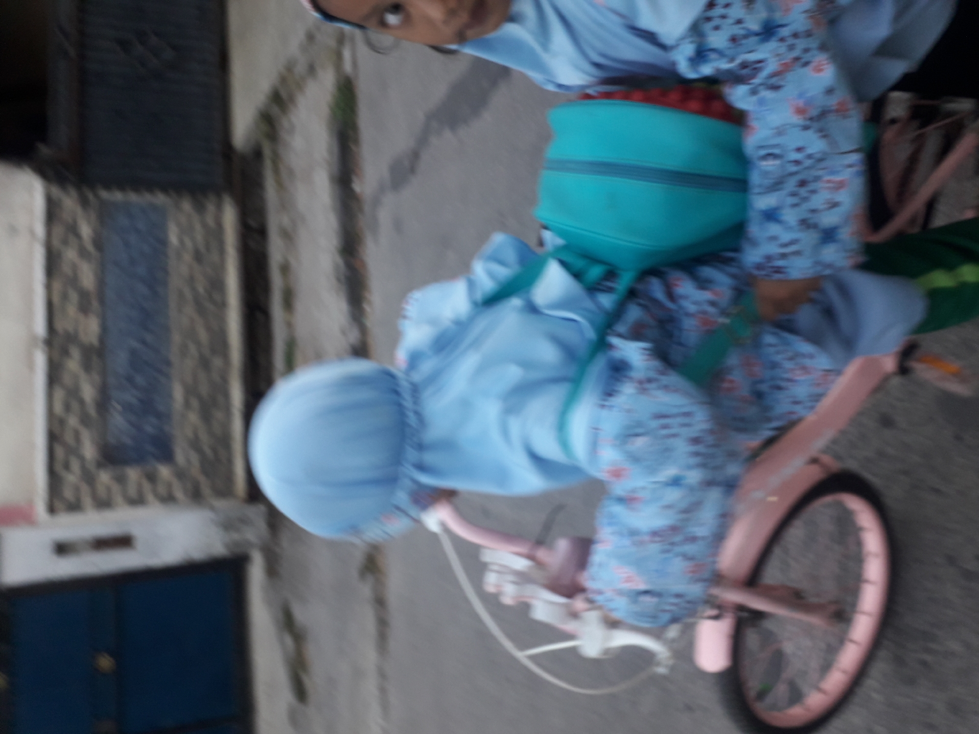 In Cangkang, Indonesia, two young girls are on a bicycle. The girl in the front is wearing a blue hijab and a blue patterned long-sleeved shirt. She is holding onto the handlebars of a pink bicycle. The girl in the back, also wearing a blue hijab, is sitting behind the first girl and has a turquoise bag strapped to her. Her face is visible to the right of the frame, and she appears to be looking at the camera. They are on a paved road, with a blurred background of buildings and a wall. The image has a motion blur effect, suggesting the bicycle is moving.