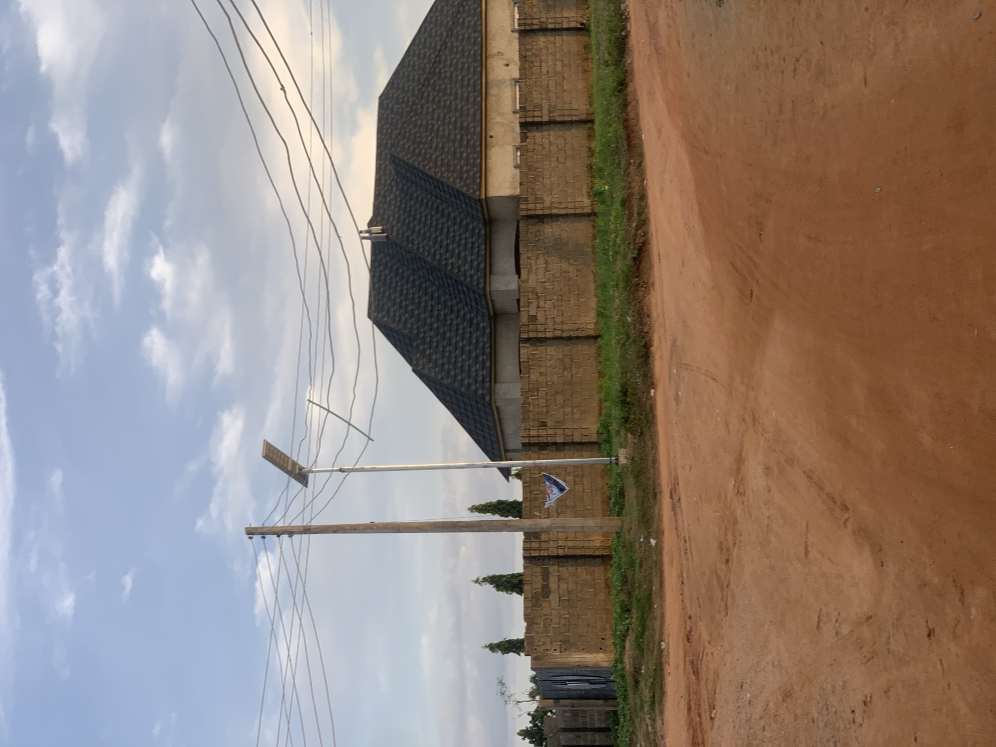In Rido, Nigeria, a dirt road curves through a developing area under a partly cloudy sky. A low, cinderblock wall encloses a property, topped by a dark, asphalt-shingled roof of a single-story building. The house appears unfinished, with visible window openings. A wooden utility pole stands beside the road, supporting multiple power lines. A solar-powered street light is mounted on a metal pole near the wooden one. A small, blue and white flag or banner is attached to the solar light pole. Several tall, conical evergreen trees are visible behind the wall, along with other fencing. The foreground is dominated by the reddish-brown dirt road, showing tire tracks and scattered gravel. The overall impression is one of a semi-rural or suburban environment undergoing construction.