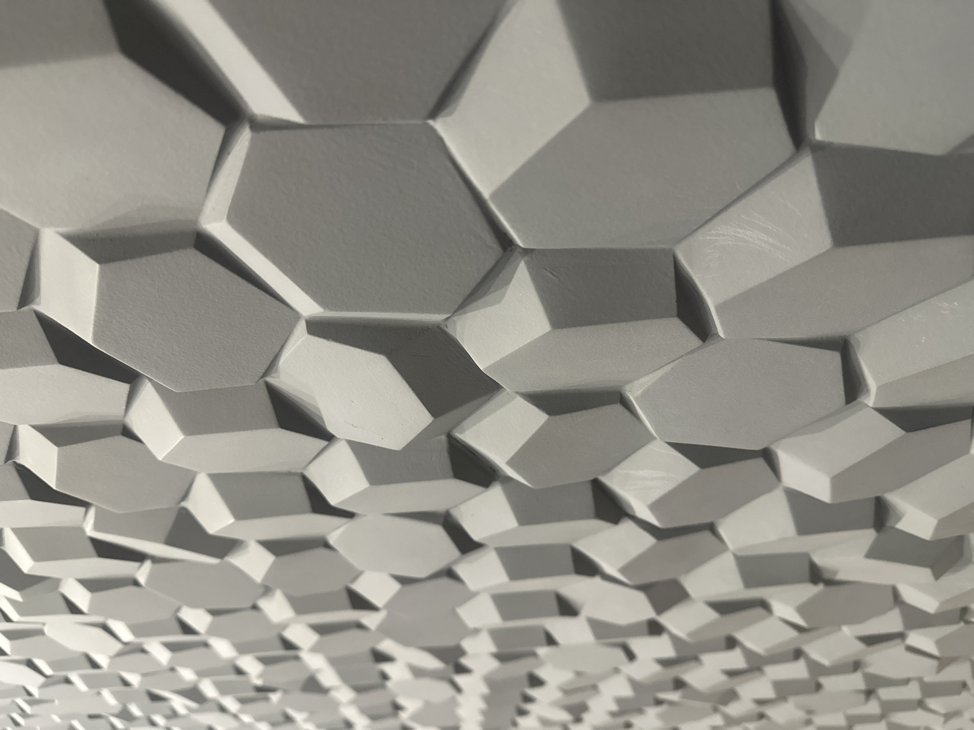 The image presents a close-up, angled view of a textured architectural wall surface in Izhevsk, Russia. The surface is composed of numerous irregular, three-dimensional geometric protrusions, primarily resembling faceted hexagons and pentagons. These elements are tightly tessellated, creating a continuous, relief pattern that extends across the visible area. Each shape projects outward, exhibiting beveled edges and varied depths. The entire surface is rendered in a uniform light gray, matte tone. Strong directional lighting from the upper left casts prominent shadows beneath and to the right of each raised element, accentuating the three-dimensional relief and creating depth. The perspective is from the left, looking diagonally across the surface, with elements in the foreground (right side) appearing larger and more in focus, gradually diminishing in size and clarity towards the background (left side) due to shallow depth of field. Subtle surface imperfections and fine scratches are visible on some facets, indicating a physical, possibly sculpted or molded material. No individuals, specific actions, or other identifiable objects are present in the scene.