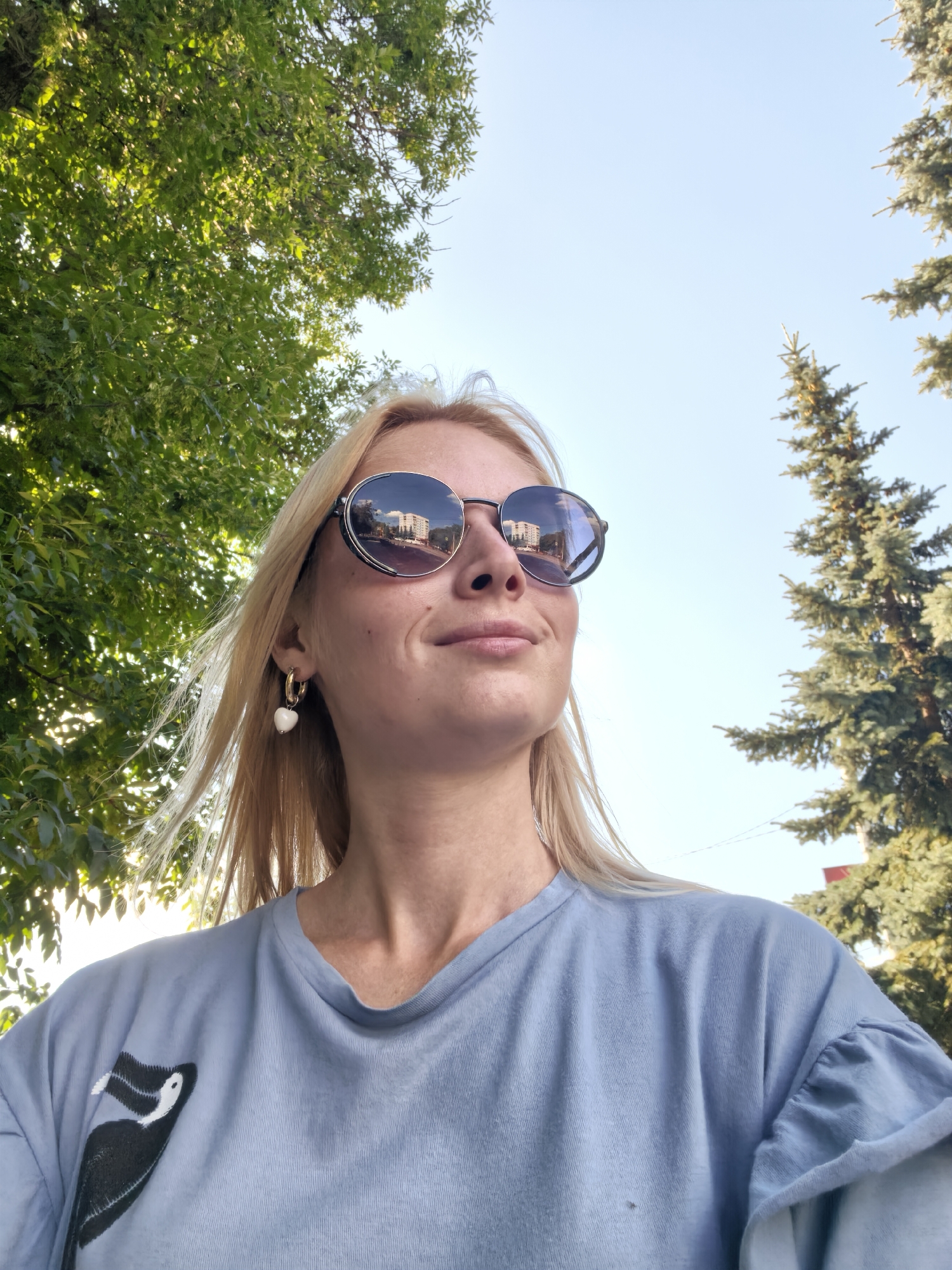 A medium-shot, slightly low-angle, outdoor selfie shows a blonde woman with fair skin wearing a light blue t-shirt with a black and white cartoon bird graphic on the left chest. She is wearing round, oversized sunglasses with a dark rim and reflective lenses that show a reflection of buildings and a street. She has a white heart-shaped earring on her left ear. Her hair is styled straight and falls over her shoulders. The background consists of a bright blue sky with sunlit green leaves from a tree on the left and a tall, dark green evergreen tree on the right. This scene is consistent with a sunny day in a park or residential area of Sterlitamak, Russia.