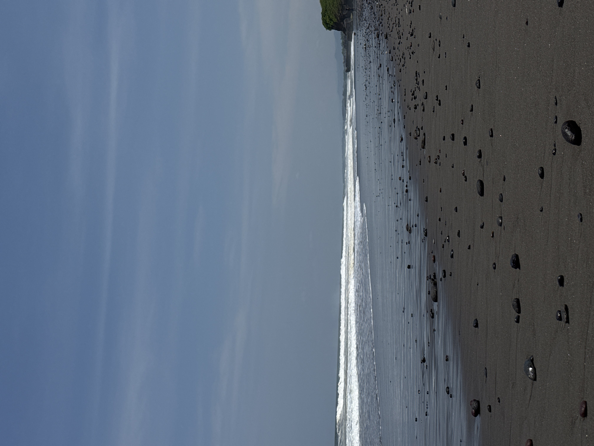 The image depicts a volcanic black sand beach in Sudimara, Indonesia, under a clear to partly cloudy daytime sky. The upper third of the frame shows a pale blue sky with minimal, high-altitude cloud formations. Below the horizon, the ocean extends across the mid-ground, featuring several lines of breaking white-capped waves receding towards the shore, with the water appearing dark and reflecting the sky near the wet sand. The foreground and lower mid-ground are composed of dark, fine-grained sand heavily scattered with numerous smooth, dark, irregularly shaped pebbles and small rocks. The sand is visibly wet and reflective closest to the water, gradually becoming drier further up the beach. On the far right, a vegetated rocky headland or cliff partially obstructs the distant horizon, with faint outlines of further landforms visible beyond it. No identifiable individuals or specific human activities are present within the depicted scene.
