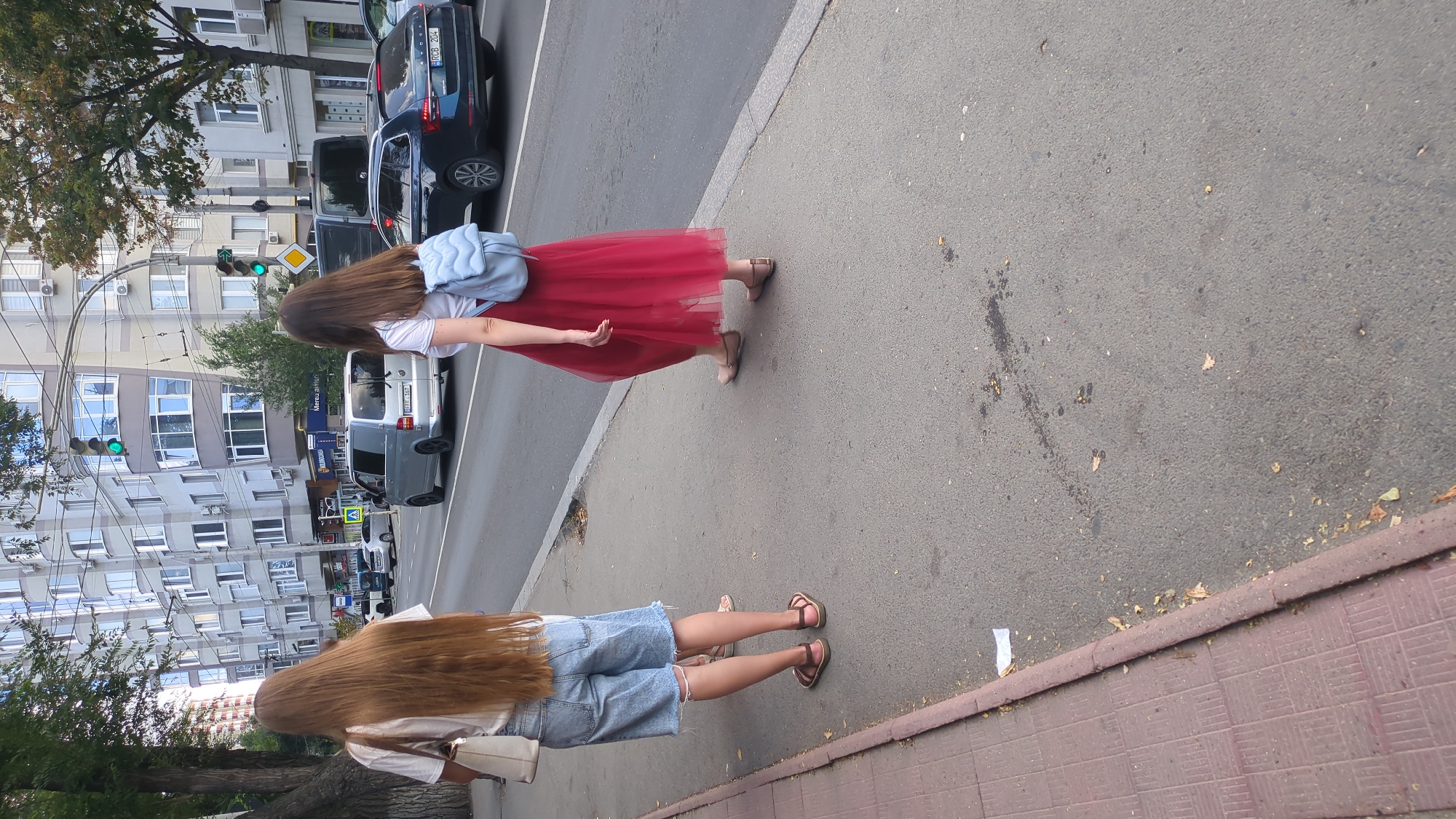 Two young women are walking on a sidewalk next to a city street in Chișinău, Moldova. The woman on the left has very long, light brown hair and is wearing a white t-shirt, denim shorts, and brown sandals. She is carrying a cream-colored tote bag. The woman on the right has dark brown hair and is wearing a white t-shirt, a long, flowing red skirt, and flat beige sandals. She has a light blue backpack on.
The street is busy with traffic. A silver van and a dark blue station wagon are visible. In the background, there are multi-story buildings with many windows, characteristic of urban architecture. Traffic lights are visible on poles above the street. Trees line the sidewalk and street. The sidewalk is paved with red tiles on the left and asphalt on the right. Fallen leaves are scattered on the sidewalk and street. The scene appears to be in the daytime.