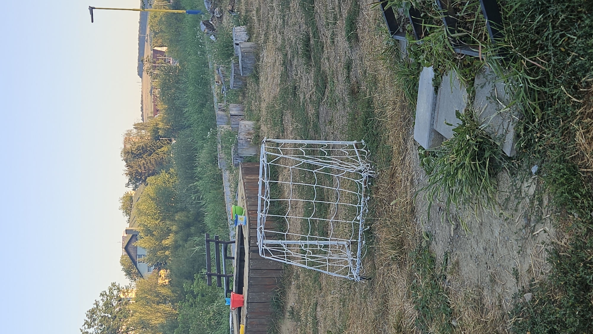 The image depicts an outdoor area, likely a park or playground, with a small, portable soccer goal in the foreground. The goal is white with white netting and appears to be made of metal tubing. Behind the goal, a wooden sandbox with red and green buckets is visible, along with a dark wooden bench. The ground is covered in dry grass and dirt. In the background, there are trees, some houses, and a yellow and blue lamppost. The overall scene suggests a somewhat neglected or underutilized recreational space. The image is taken during daylight hours, under a clear sky. No people are present in the image. The location is identified as Prešov, Slovakia.