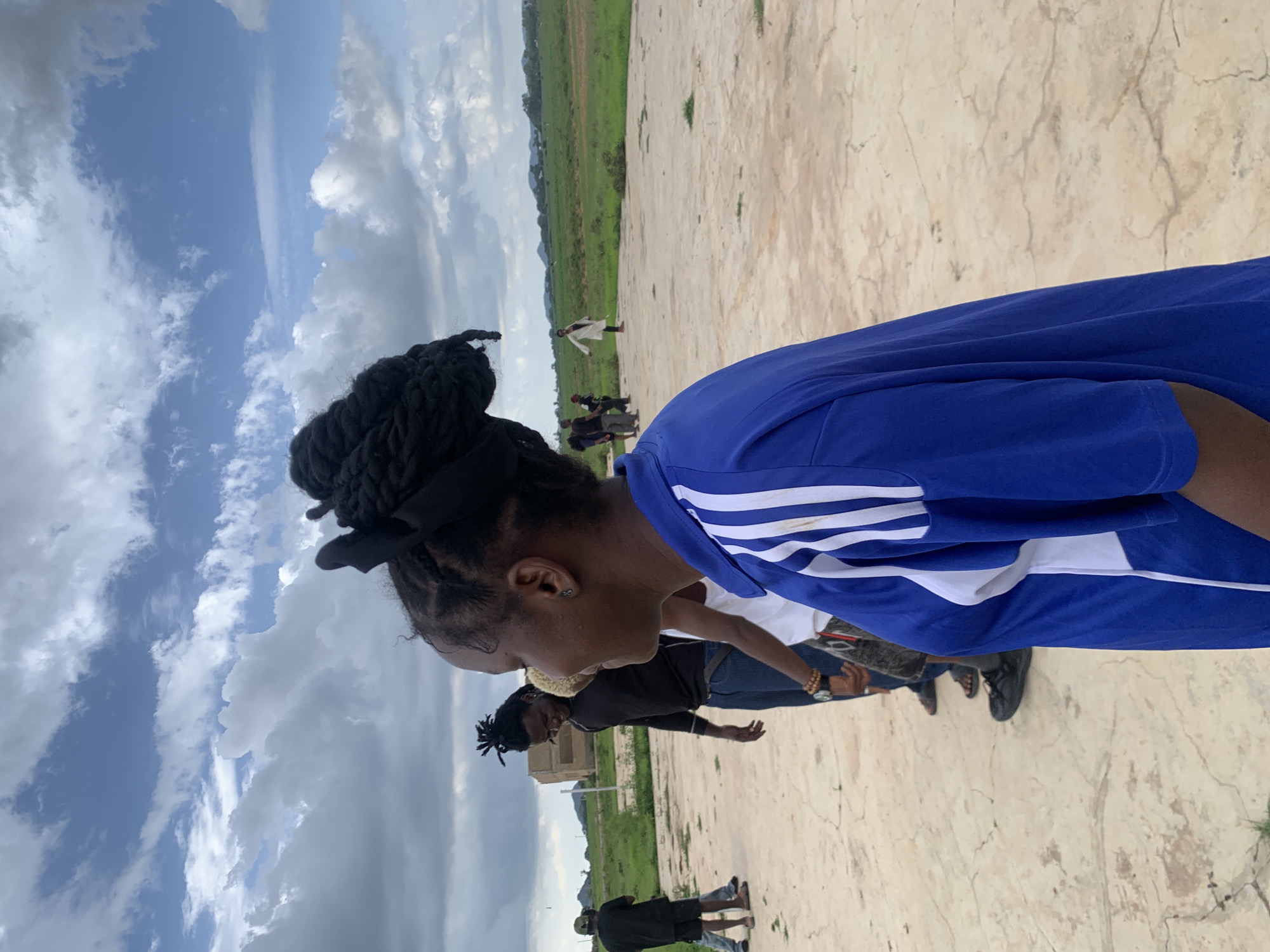 The image depicts a group of people outdoors in Gwafan, Nigeria. The scene appears to be a wide, open, paved area with a grassy field and some distant buildings in the background. The sky is partly cloudy with dramatic, billowing clouds against a bright blue sky, suggesting it might be daytime, possibly late morning or afternoon.
In the foreground, a young woman is captured from the side, her head bowed and turned to her left. She has dark hair styled in braids, tied up with a black ribbon. She is wearing a vibrant blue t-shirt with white stripes on the sleeves. Her expression is not clearly visible, but her posture suggests focus or contemplation.
Several other individuals are visible in the mid-ground and background, some appearing to be engaged in activities or standing around. One person in the background is wearing a white garment. Another group of people further back includes individuals in dark clothing, and one person in a white shirt and blue jeans. The ground beneath them is a light-colored, textured surface, possibly concrete or stone.
The overall atmosphere seems relaxed and casual, with people gathered in an outdoor space. The weather appears to be pleasant, with the dynamic clouds adding visual interest to the sky. There is no visible text in the image.