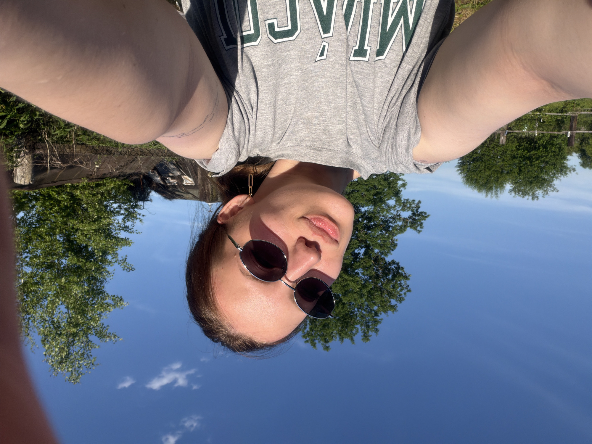 A young female is centered in a selfie taken outdoors in Reshetnikovo, Russia, under a bright blue sky with minimal cirrus clouds. She wears dark oval sunglasses with a thin metallic frame and a grey t-shirt featuring green "MIAMI" lettering. Her head is slightly tilted to her right, and her lips are slightly parted. A gold-colored chain-link earring is visible on her right ear. Faint, abstract linear markings are visible on her right upper arm. Both of her arms are extended forward, holding the camera, and are visible in the lower portion of the frame. The background consists of dense green deciduous trees, a rustic wooden fence on the left, and a dilapidated outbuilding with a dark roof and light-colored walls, partially obscured by overgrown vegetation and a wire fence on the right. The scene is illuminated by direct sunlight, casting shadows.