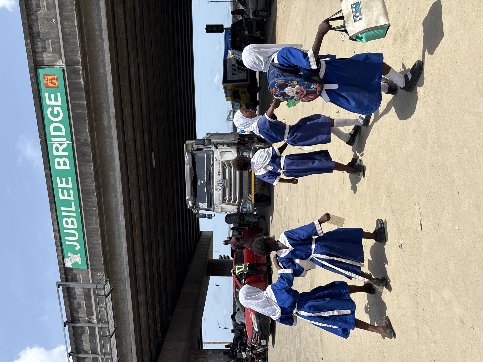 Four girls in blue school uniforms with white headscarves walk along a street under the Jubilee Bridge in Ado, Nigeria. The girls walk in a line, with one carrying a backpack and another carrying a tote bag. Behind them, there is traffic with cars and trucks on the road. The bridge sign is green with white lettering. The sky is partially visible.