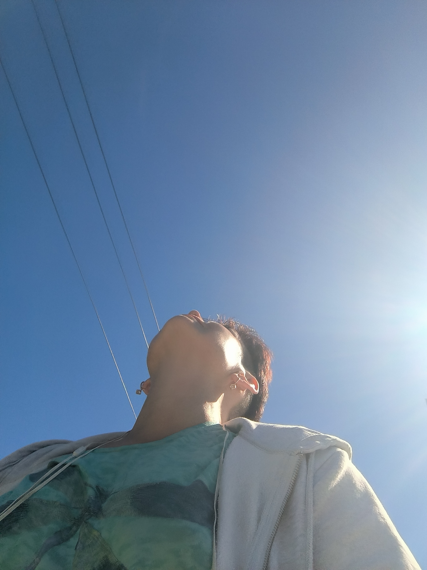A low-angle, close-up shot captures a person looking upwards into a clear, bright blue sky on a sunny day. The sun is prominently visible on the right side of the frame, casting a bright glare. Three dark, parallel lines, likely power lines, extend diagonally from the upper left corner of the image into the sky.
The person, whose face is partially obscured as they tilt their head back, appears to be wearing a teal-colored t-shirt with a dark graphic print, possibly a palm tree. They are also wearing a light-colored hooded jacket over the t-shirt. Visible jewelry includes earrings on both ears and a ring on one finger. The person's hair is a reddish-brown hue.
The overall impression is one of outdoor activity, possibly in a park or open area, given the clear sky and power lines. The bright sunlight suggests it is daytime, likely late morning or afternoon. The mood conveyed is one of peacefulness and enjoyment of the outdoors. No text is visible in the image.