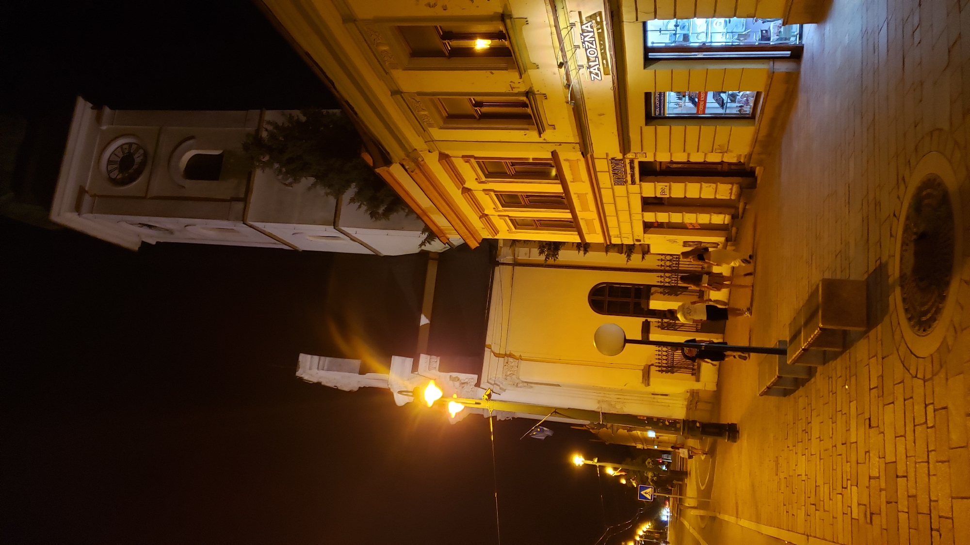 The image depicts a nighttime street scene in Prešov, Slovakia, focusing on the intersection of Slovakian culture and urban life. In the background, a prominent clock tower, likely part of a historic church, stands tall against the dark sky. The tower features a clock face with Roman numerals and a pointed spire. Adjacent to the tower, a building with white architectural details and arched windows suggests a religious or institutional structure.
In the foreground, a row of yellow buildings lines the street, their facades illuminated by streetlights and internal lighting. One building has a sign that reads "ZÁLOŽNÁ," indicating a financial institution. Another sign above a shop reads "BYZANT." The sidewalk is paved with brickwork and features several concrete benches. A lamppost with a large spherical light fixture stands near the benches.
Several individuals are walking along the sidewalk. A group of four young people, two women and two men, are walking away from the camera. Another person is walking towards the camera in the distance. The street itself is visible with traffic lights and a pedestrian crossing sign. The overall atmosphere is one of a quiet evening in a European city, with historical architecture and modern life coexisting.