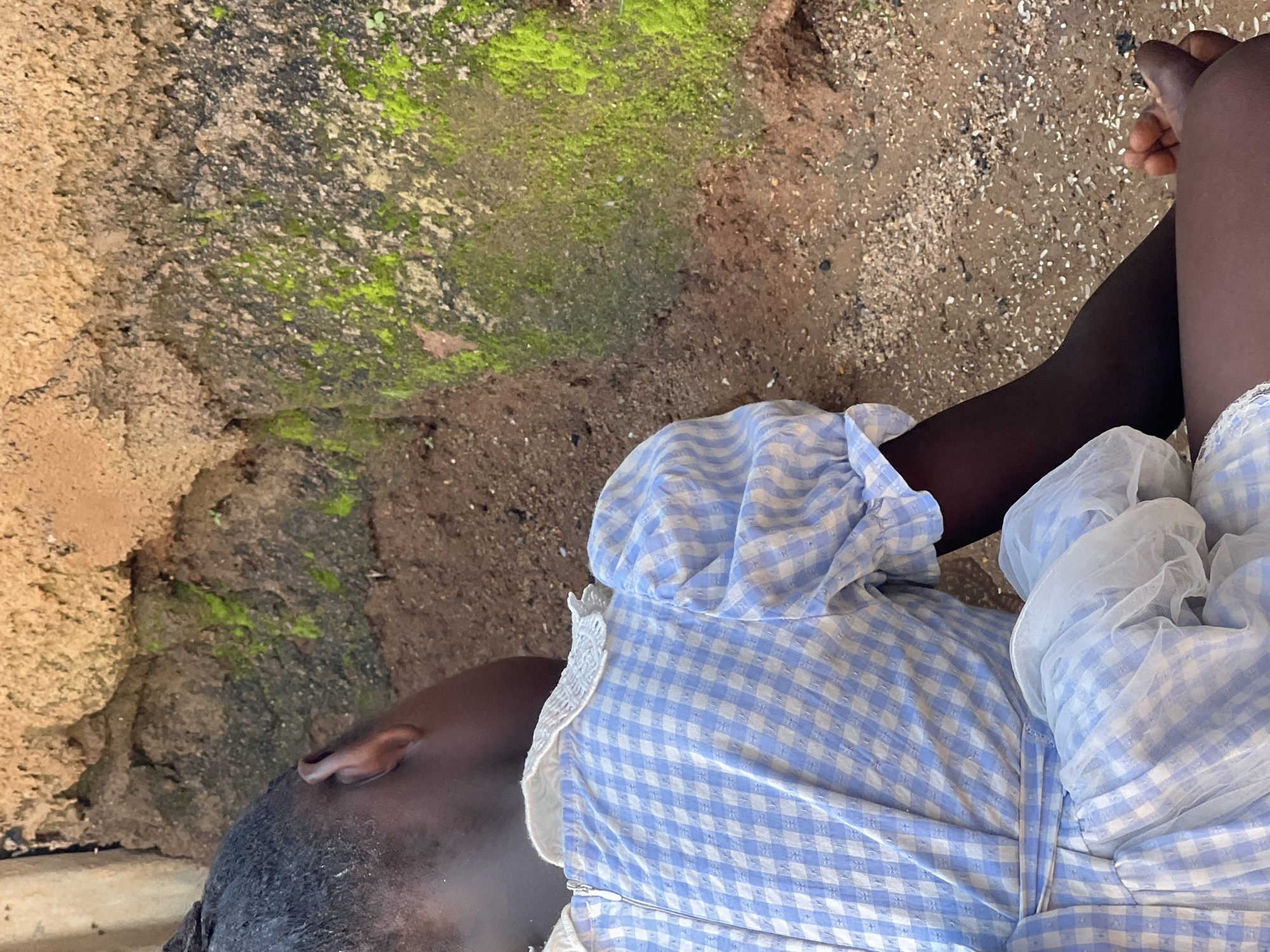 A child, seen from the back and right side, is wearing a blue and white checkered dress with white lace trim and puffed sleeves, sitting on the ground next to a rough, earthy wall. Patches of green moss grow on the lower section of the wall. The ground is composed of dirt, small stones, and some dark debris. The overall setting appears to be an outdoor area in Jos, Nigeria, characterized by rustic construction and natural elements.