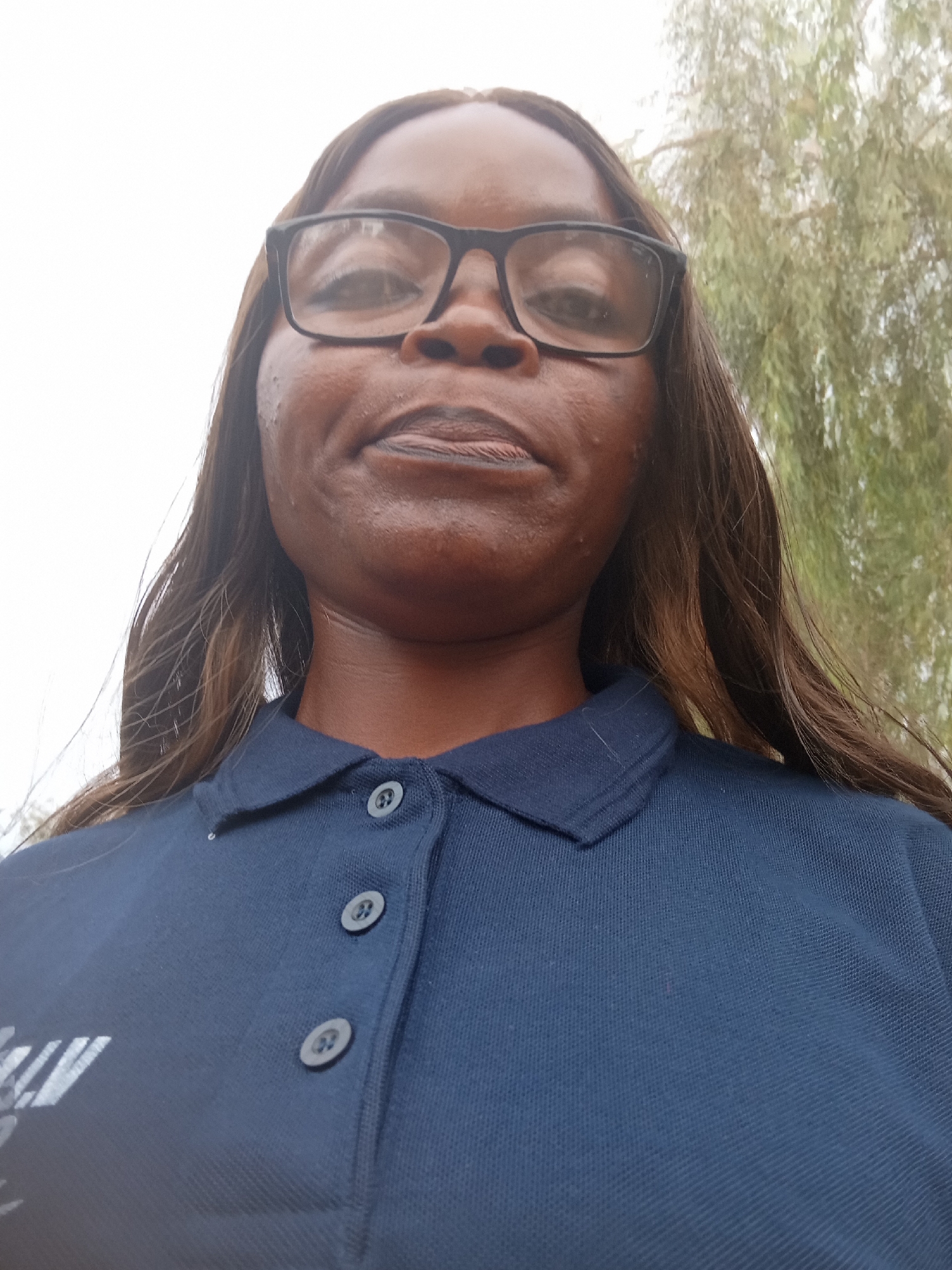 A head-and-shoulders, low-angle, outdoor, selfie shows a Black woman wearing a dark blue polo shirt and black-rimmed glasses. The woman's hair is long and brown, and she is looking at the camera. In the background, there are out-of-focus trees and a light-colored sky. The image was taken at Mayo Dasa, Nigeria.