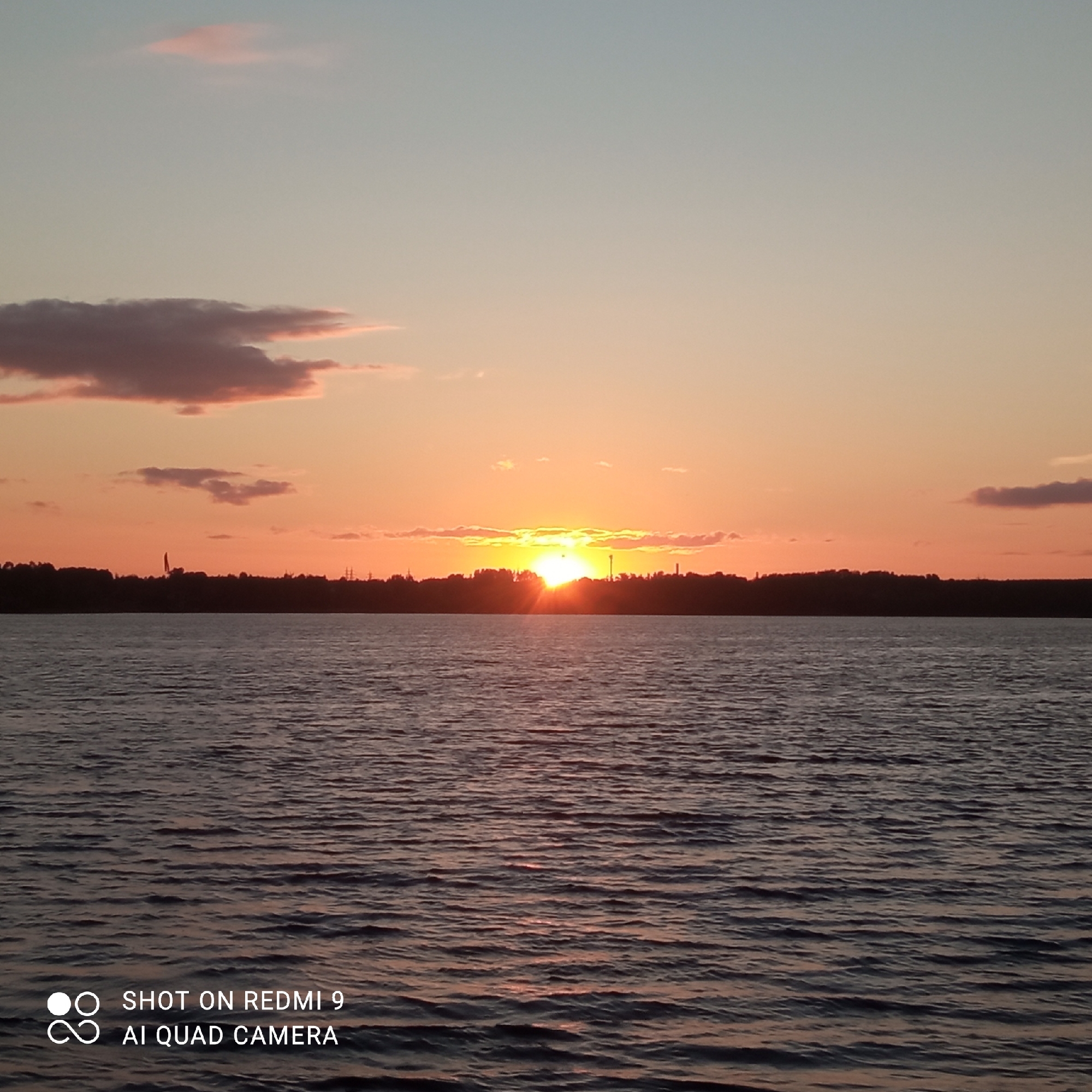 A wide-angle photograph captures a serene sunset over the Kama River in Chaikovsky, Russia. The sky transitions from a pale blue at the top to warm orange hues near the horizon, with scattered clouds tinged pink and purple. The sun, a brilliant yellow orb, is partially obscured by a dark, silhouetted treeline that stretches across the horizon. The river in the foreground is calm, with gentle ripples reflecting the colors of the sunset. The water appears dark and deep. In the lower left corner, a white watermark reads "SHOT ON REDMI 9 AI QUAD CAMERA".