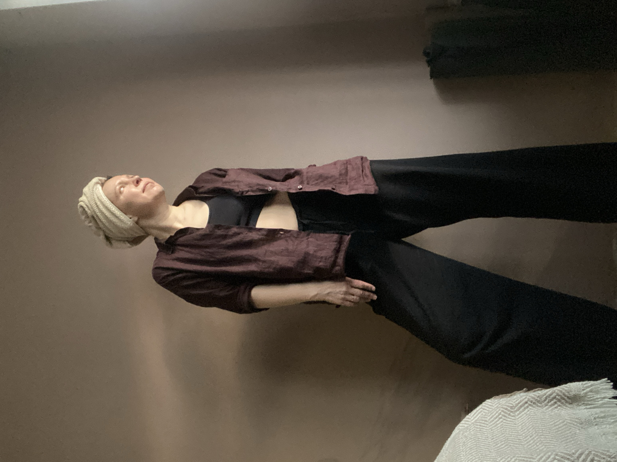 A person wearing a white knitted head covering, a black top, a maroon button-down shirt, and wide-legged black pants is standing in front of a wall with a dark beige color. A white textured blanket is visible in the bottom right corner. The location is not identifiable as Chaikovsky, Russia.