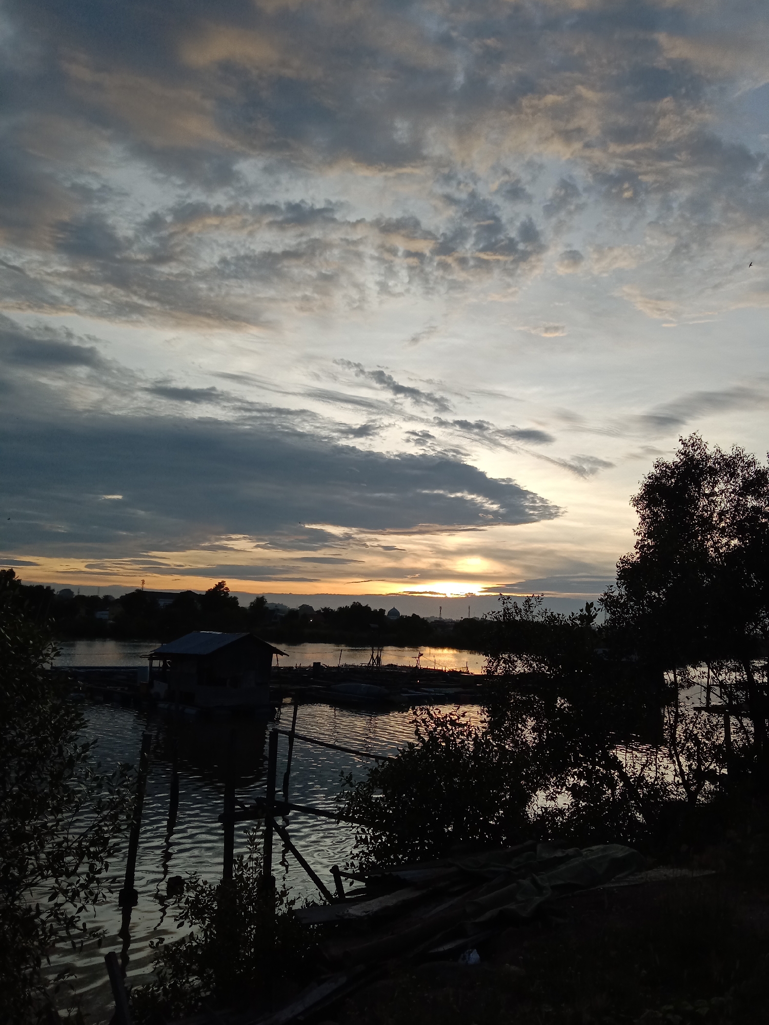 The image captures a sunset over a body of water, likely a river or bay, in Lhokseumawe, Indonesia. A small shack is built on stilts in the water, with wooden structures and posts supporting it. The water reflects the orange and yellow hues of the setting sun. In the background, a silhouette of the city skyline is visible, including a mosque dome and possibly other buildings. The sky is filled with varied cloud formations, ranging from dark gray to light gray and white. The foreground is dominated by dark foliage, including trees and bushes, partially obscuring the view of the water.