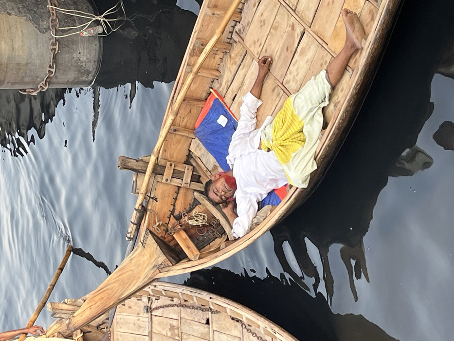 The image captures a scene on dark water, consistent with a riverfront or dock setting in Dhaka, Bangladesh, possibly along the Buriganga River. In the foreground, an adult male lies supine within a wooden boat. He is dressed in a white long-sleeved shirt and a light yellow and white striped lower garment, possibly a lungi. A significant quantity of a red substance, appearing consistent with blood or a similar thick fluid, is smeared across his chin, mouth, and the left side of his face. His eyes are closed, and he appears unresponsive. His head rests on a folded blue and red fabric item. His left arm is bent, with his hand near his head, while his right arm is extended along his side. The boat is a traditional wooden vessel, featuring plank construction and an oar or wooden pole resting inside along the starboard side. To the left, a portion of another similar wooden boat is visible, showing a metal chain attached to its bow. In the upper right background, a concrete cylindrical pier or mooring post rises from the water, encircled by a rusty chain, with a plastic bottle tied to it by a string. The dark water reflects the objects and sky.