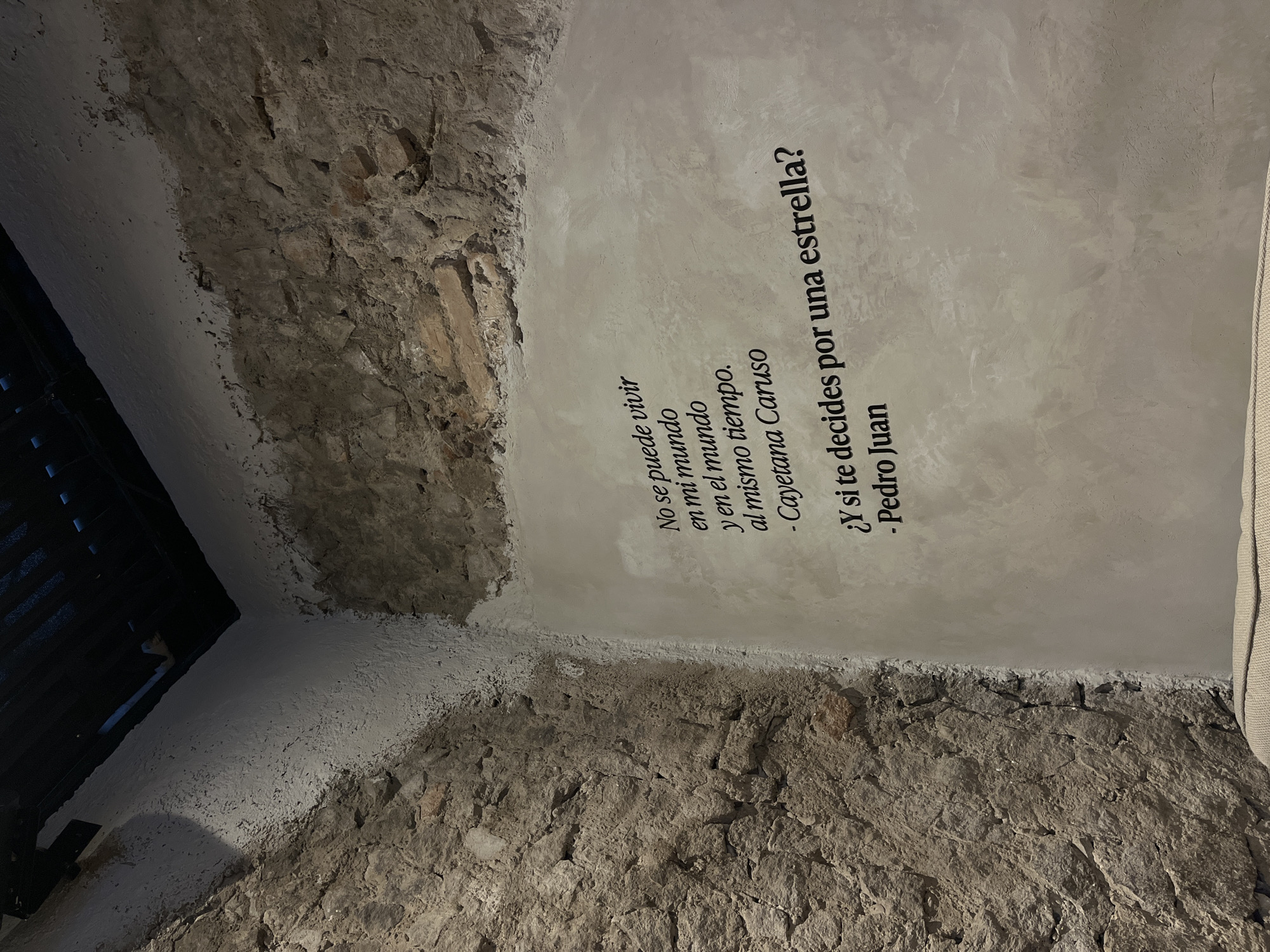 The image presents a corner of a room, possibly an interior wall or ceiling, where modern construction meets exposed stonework. The stonework, likely part of the building's original structure, is visible along the bottom and left side of the image, showcasing the rough texture of the aged stones. The upper and right sections are plastered with a smooth, gray-toned surface, contrasting with the rustic stonework. A text overlay in Spanish is printed on the plastered surface: "No se puede vivir en mi mundo y en el mundo al mismo tiempo - Cayetana Caruso", followed by the question "Y si te decides por una estrella? - Pedro Juan". This suggests a philosophical or poetic theme to the space. A partial view of what appears to be a dark, slatted window or gate is visible on the extreme left, and the edge of a light-colored piece of furniture or fixture is seen on the extreme right. The setting is likely indoors, in a building that blends old architecture with modern elements. The city of San Bernardino Tlaxcalancingo, Mexico is mentioned as the possible location of this place.