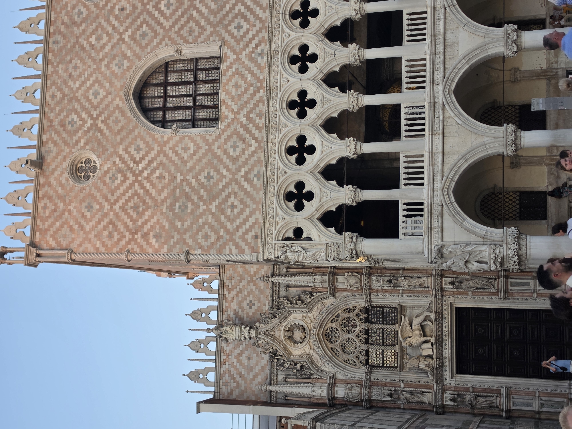 The image shows a detailed view of the Doge's Palace in Venice, Italy. The architecture is Gothic, with intricate stonework, arched windows, and decorative carvings. The facade features a diamond pattern of pink and white marble. In the foreground, a dark wooden door is visible, flanked by statues and ornate carvings. Above the door, a relief depicts a winged lion with a book. To the right, a loggia with a series of quatrefoil windows is supported by columns. The sky above is clear and pale blue, suggesting it is either early morning or late afternoon. Several people are visible in the lower part of the image, some looking at the camera, others appearing to take photographs. A person in a light blue shirt is standing in front of the door, holding up a phone. The overall scene captures the grandeur and historical significance of the Venetian landmark.