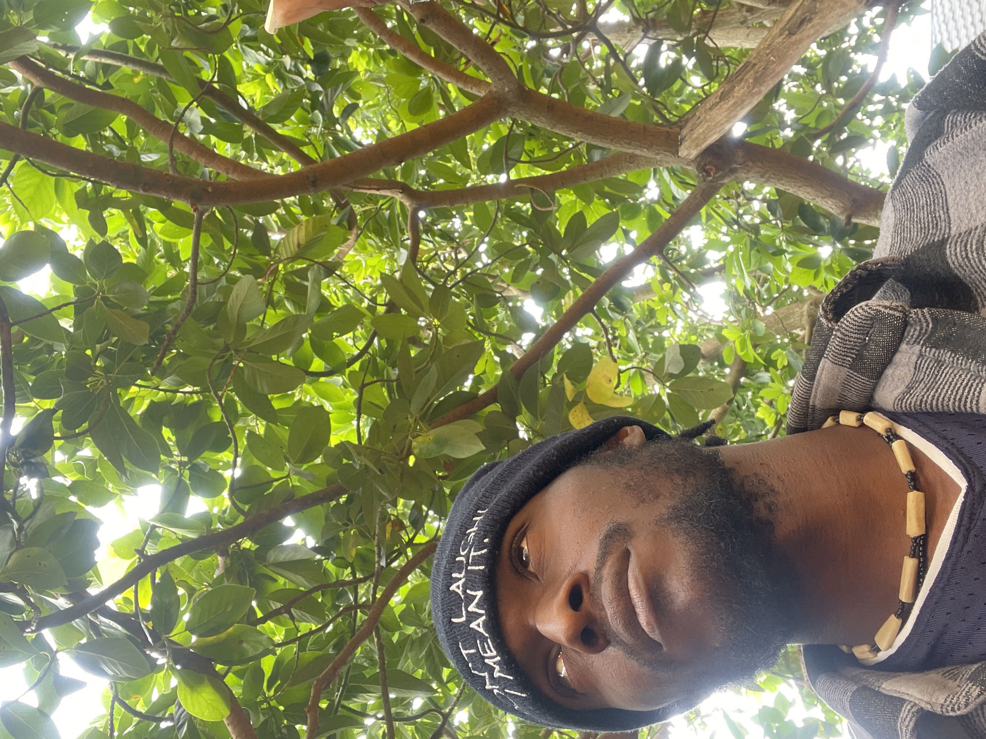 A low-angle, color photograph depicts a Black man in Jos, Nigeria, standing under a tree. The man is wearing a black beanie with white lettering that reads "LAUGH LIKE I MEAN IT," a necklace of beige and black beads, and a blue jersey under a gray and black plaid jacket. The background is filled with green leaves and brown branches of the tree, with the sky peeking through in places. The man is looking down at the camera.