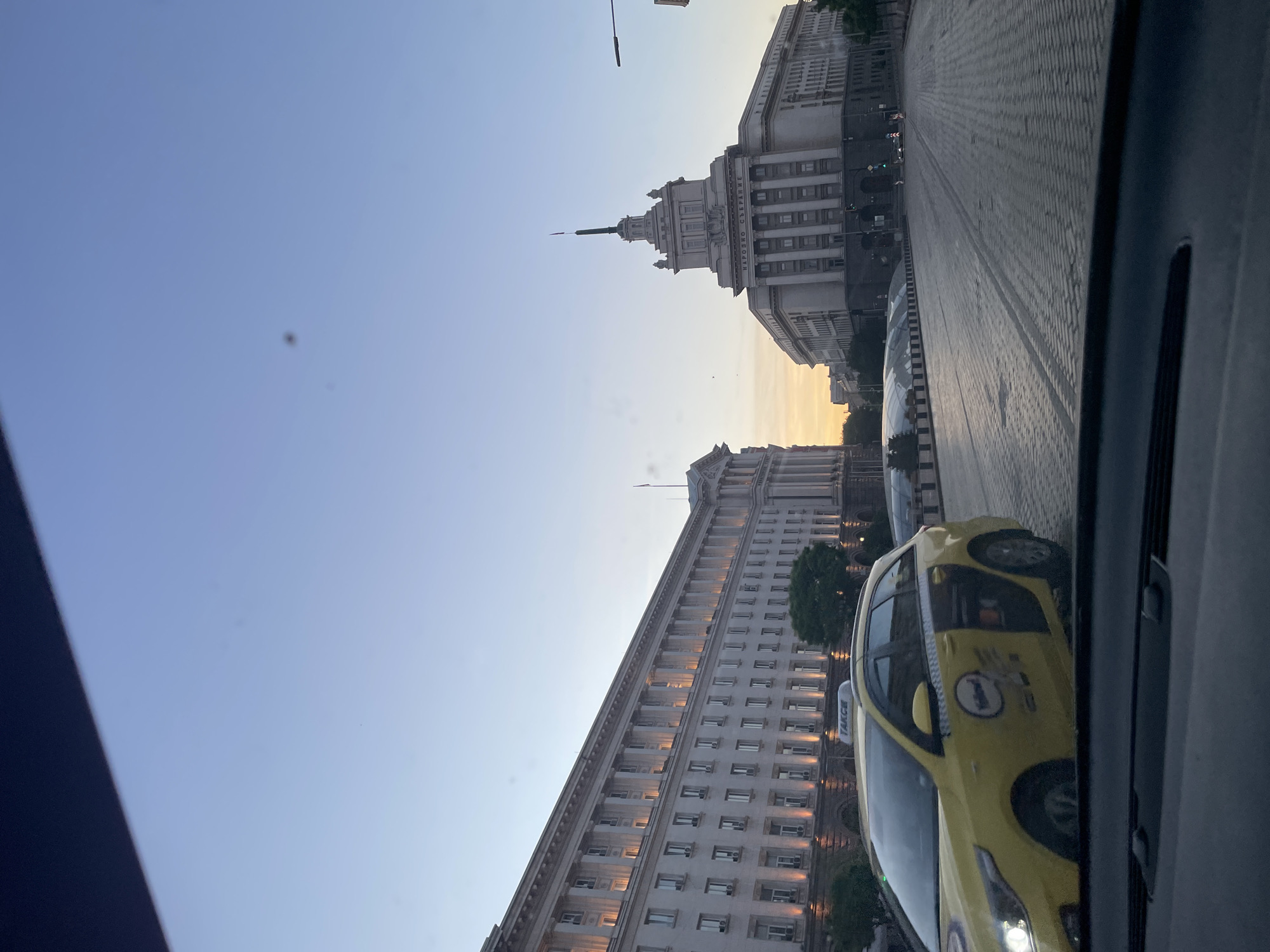The image shows a view from inside a car in Sofia, Bulgaria, looking towards the Largo. Visible are parts of the National Assembly building (with its distinctive spire), the Presidency building, and the Council of Ministers building. The street appears to be largely empty. A yellow taxi is partially visible in the foreground, on the right. The sky is a gradient of blue and orange, suggesting either sunrise or sunset.