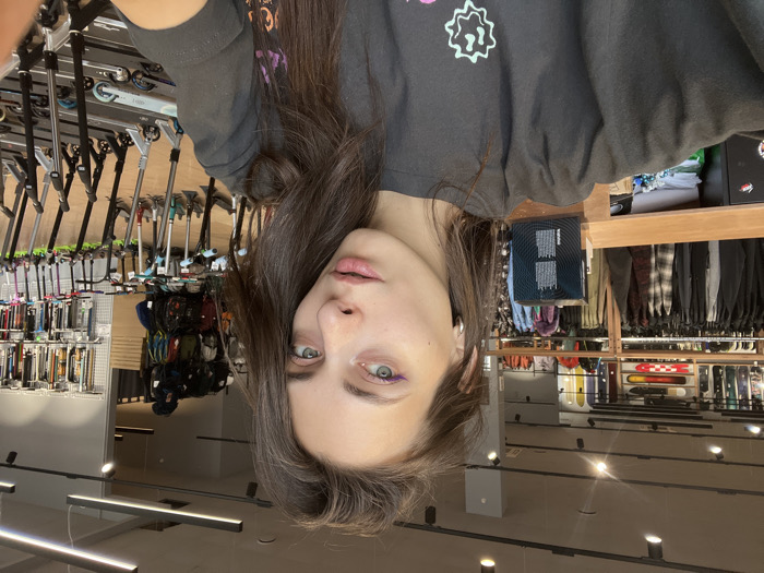 A female individual is depicted in a head-and-shoulder selfie captured within a retail store in Krasnoyarsk, Russia. She has long, dark brown hair, light-colored eyes accented with purple eyeliner, and wears a dark-colored short-sleeved t-shirt with a light graphic design on the left chest. A silver hoop earring is visible on her left ear. Her mouth is slightly ajar.
The background reveals a commercial interior. To the subject's left, a clothing display features dark garments on racks, with multiple snowboards or skateboards mounted horizontally on a wall above. To the subject's right, numerous kick scooters or stunt scooters are arranged in rows on the floor, while various backpacks and accessories hang on grid display panels above them. The store features a light-colored floor, modern track lighting, and linear LED fixtures on the ceiling. The overall environment is consistent with an action sports or streetwear retail establishment.