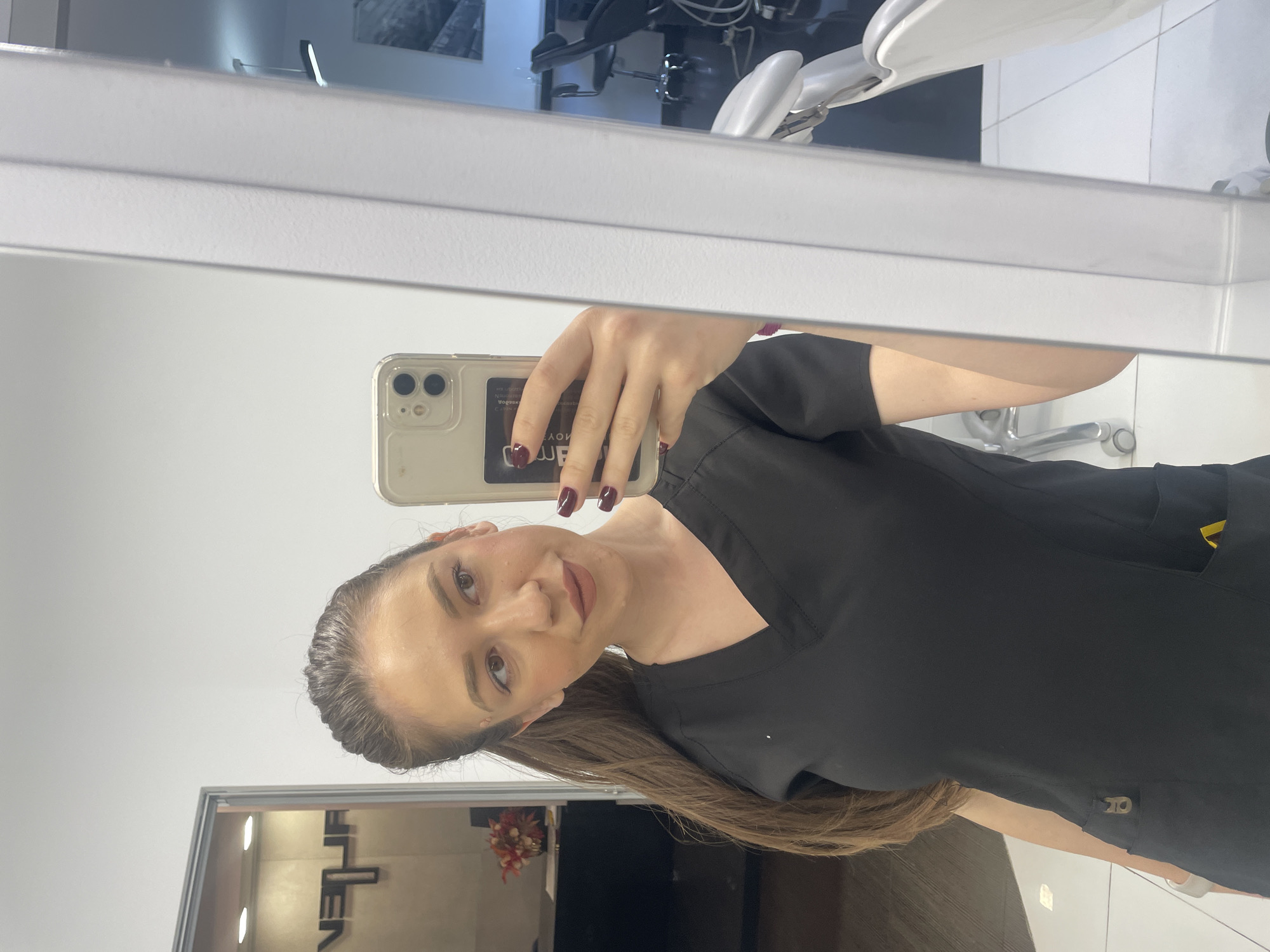 A woman is taking a selfie in a mirror inside what appears to be a dental or medical office. The woman has brown hair pulled back and is wearing a black scrub top. Her nails are painted a dark burgundy color. She is holding a smartphone with a light-colored case.
The reflection shows a dental chair and equipment, indicating a dental or medical setting. A sign is visible in a doorway, with the word "FILENA" partially visible. The location is Sofia, Bulgaria, based on the signage present in the reflection. The environment appears to be clean and modern. The woman is looking at the camera with a slight smile.