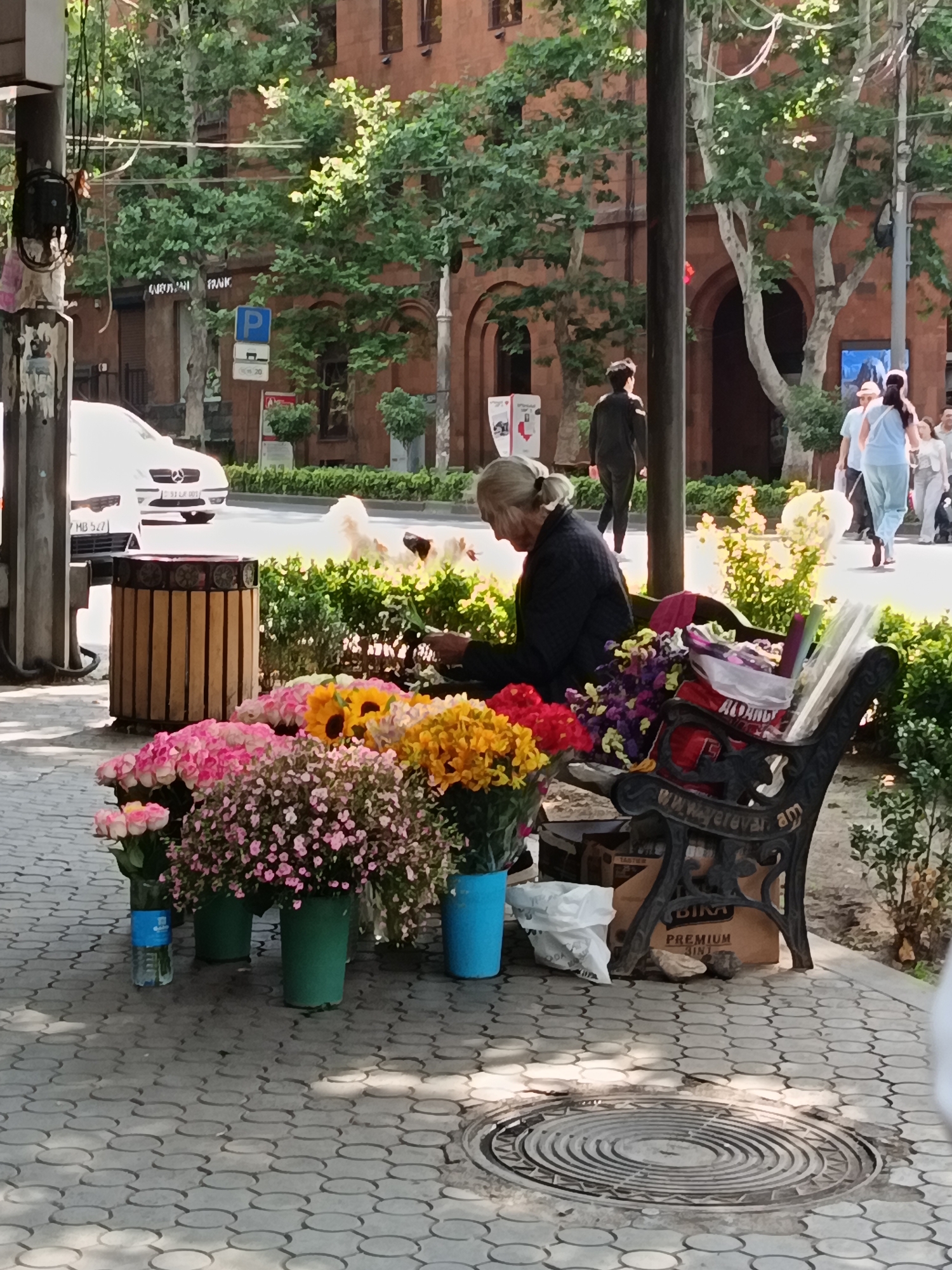 An elderly woman with gray hair tied back in a bun sits on a park bench in what appears to be an urban setting. She is wearing a dark jacket and is looking down, possibly tending to flowers. In front of her, a colorful assortment of flowers, including pink roses, sunflowers, and other blossoms, are arranged in green and blue vases. The bench itself is ornate, made of black metal with decorative patterns and inscribed text. Behind the woman, a white car is parked on a street, and people are walking by on a sidewalk. The background shows a building with red brick walls and trees with lush green foliage. The ground is paved with interlocking gray stones, and a manhole cover is visible.