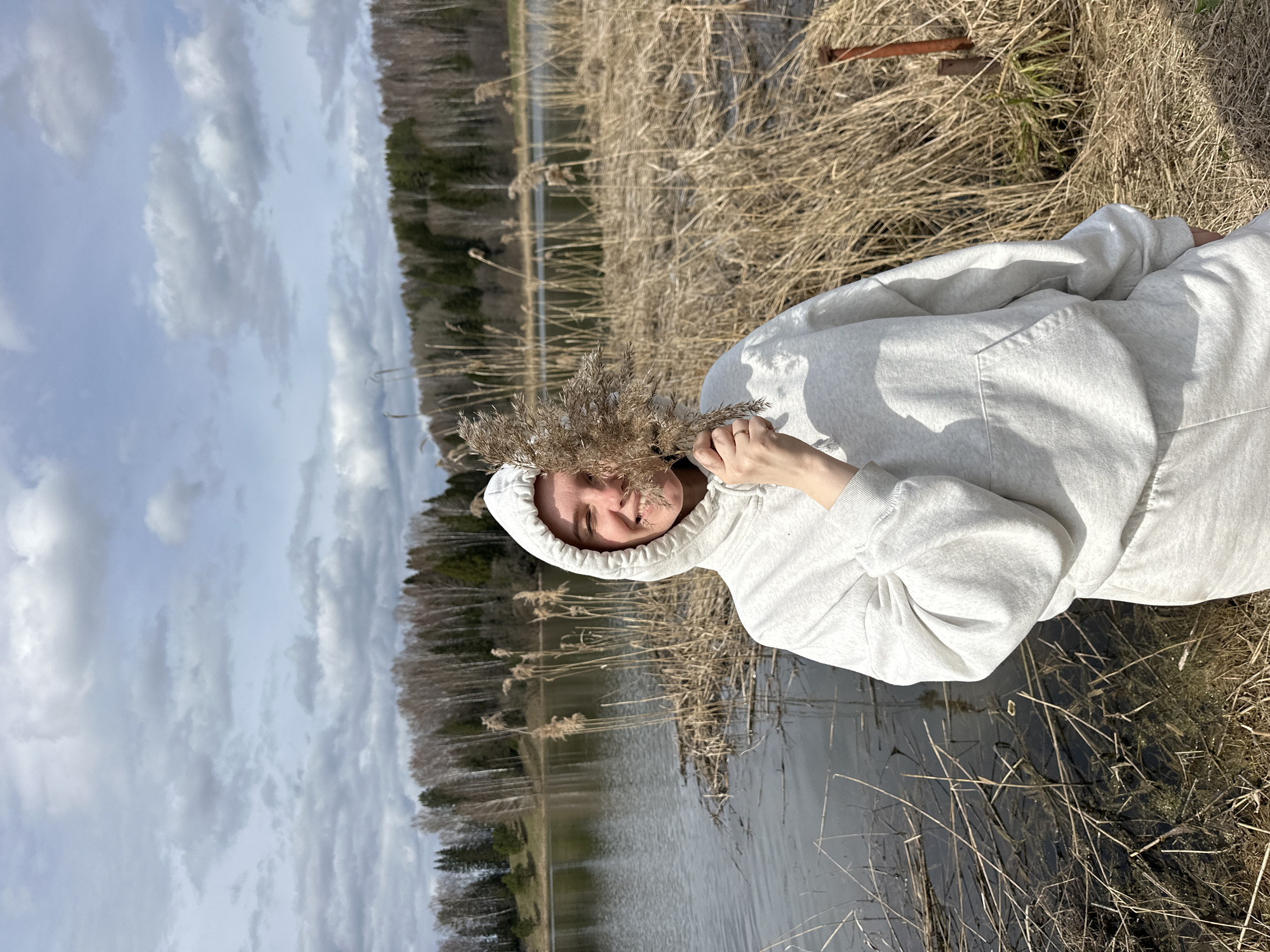 This vertically oriented image captures a person outdoors in a natural setting near a body of water. The subject, likely female, is positioned in the foreground, wearing a light grey hooded sweatshirt. They are holding a cluster of dried, light brown plant stalks (reeds or dry grass) in front of their face, partially obscuring it. A slight smile is visible around the mouth, and one eye is visible, suggesting a playful or serene expression. A simple ring is visible on the ring finger of their left hand.
The background features a tranquil body of water, possibly a lake or river, bordered by more dry, tall grass and reeds. A dense line of trees, some bare and some evergreen, forms a distant horizon under a cloudy, light blue sky. The dry nature of the vegetation suggests either late autumn, winter, or early spring. The overall lighting indicates it is daytime, under overcast conditions. In the upper right corner of the frame (when viewing the original image in landscape orientation), a piece of rusted metal is partially visible on the ground amidst the dry grass. The scene is located in Альняш, Russia. No text is visible in the image.