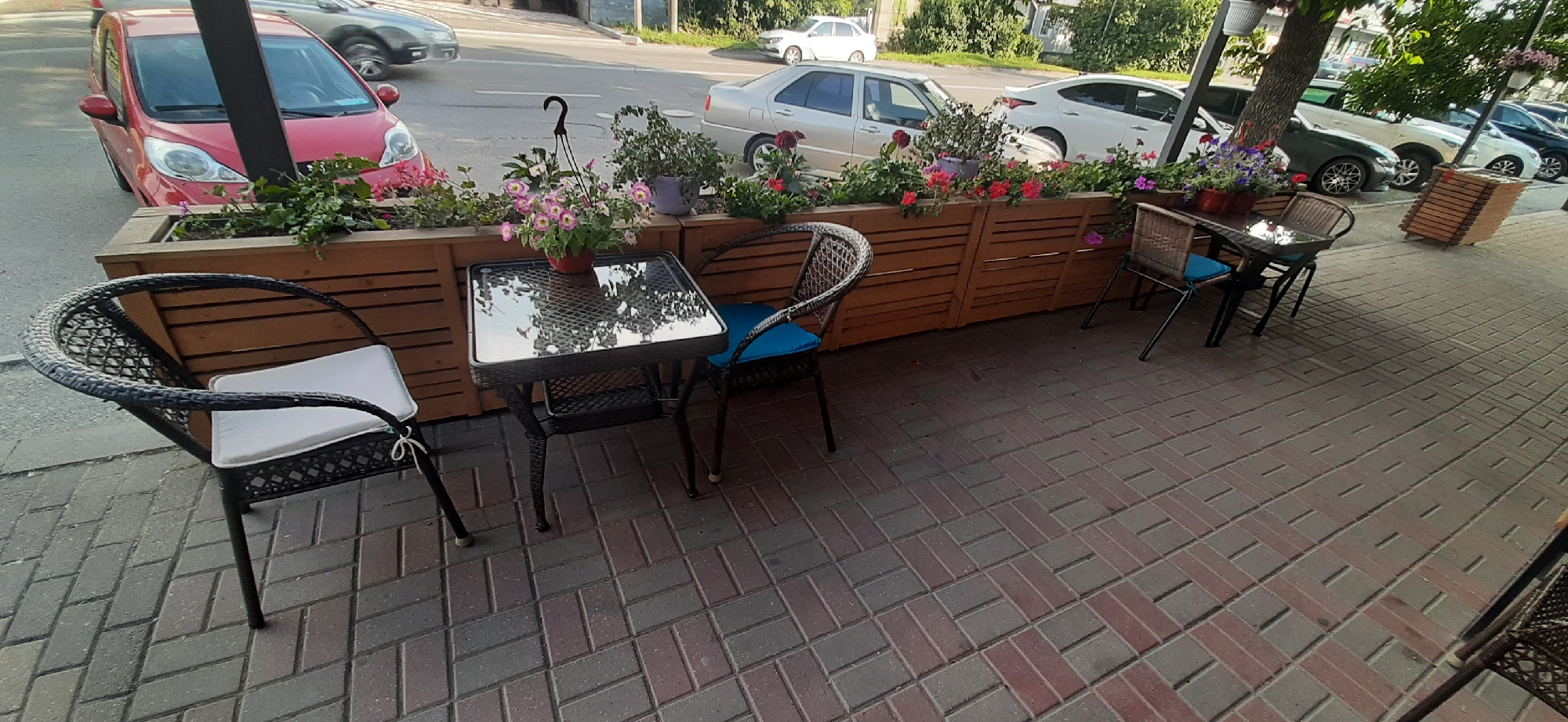 This media file shows an outdoor seating area for a cafe or restaurant, likely in Yessentuki, Russia, during the daytime. The scene is set along a paved sidewalk next to a street with several cars parked and driving by. The seating area features several tables and chairs made of woven dark material. One table has a glass top and a potted pink flowering plant hanging above it. The chairs have white cushions, and one chair also has a blue cushion.
Behind the seating area is a long wooden planter box filled with various flowers and greenery, including pink and red blooms. The planter box is made of horizontal wooden slats. In the background, cars are visible on the street, including a red car in the foreground, a silver sedan, and several other vehicles further down the road. Trees with green foliage line the street.
The lighting suggests it is daytime, with sunlight casting shadows on the ground. The weather appears to be pleasant, suitable for outdoor dining. There are no people visible in the image. The overall impression is a quiet, inviting outdoor space ready for customers. No visible text is present in the image.