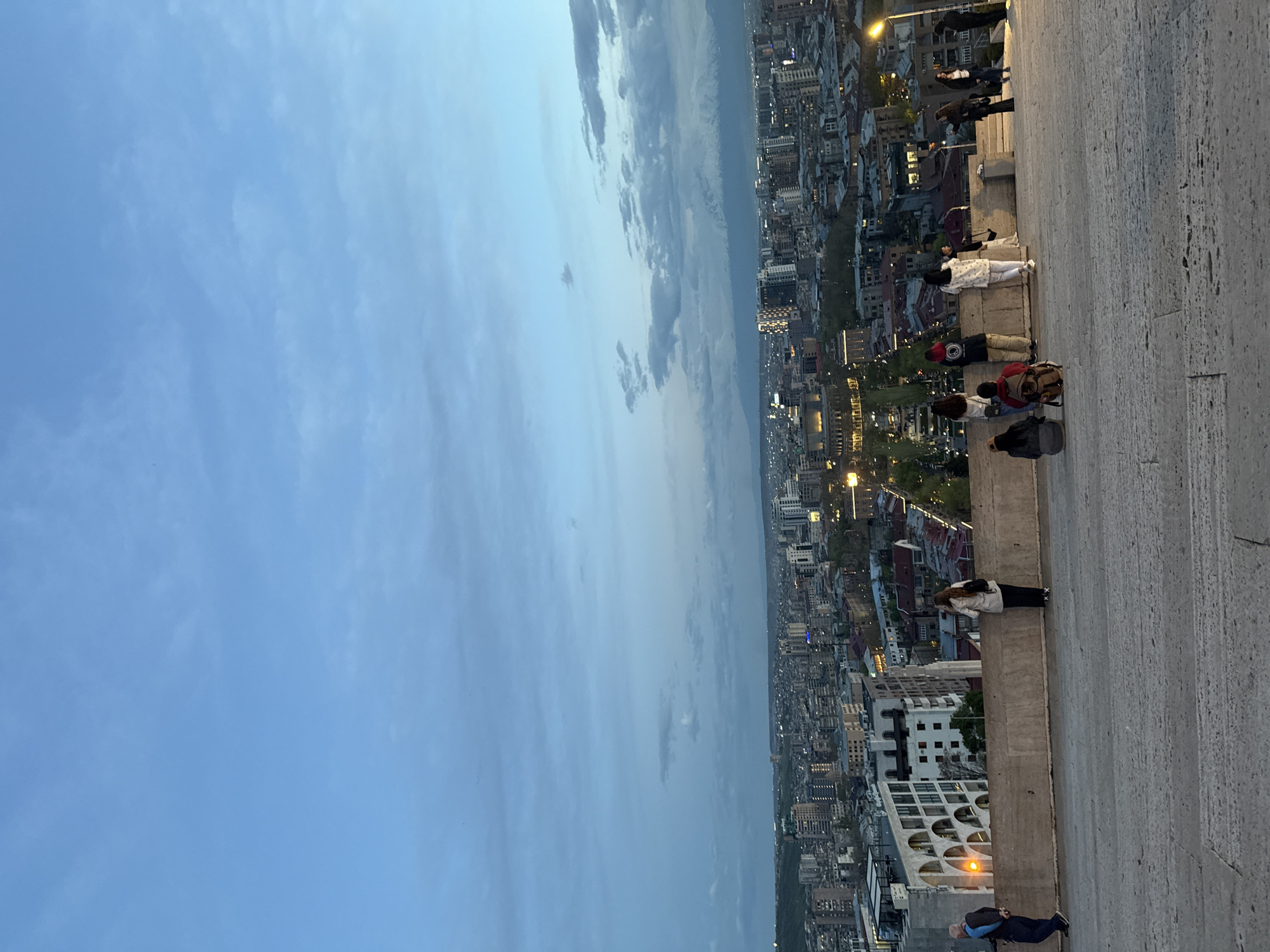 The image presents a panoramic view of Yerevan, Armenia, captured from an elevated perspective during dusk or early evening. The sky, a blend of soft blue hues with scattered, wispy clouds, dominates the upper part of the frame, gradually meeting the sprawling urban landscape below. As daylight fades, numerous city lights begin to twinkle, illuminating the diverse array of buildings that stretch across the horizon, hinting at the vastness of the city.
In the foreground, a wide, textured concrete or stone platform serves as a vantage point. Several individuals are visible on this platform; some are standing, while others are seated along the edge, all directed towards the extensive city view. They appear to be observing the urban panorama, possibly engaged in conversation, taking photographs, or simply enjoying the peaceful transition from day to night. Their faces are not clearly discernible, but their presence emphasizes the popular nature of this viewpoint. The architecture of some visible buildings, particularly one with distinctive arched windows in the mid-ground, suggests the location might be near a prominent landmark like the Yerevan Cascade. The scene conveys a serene atmosphere of appreciation for the city's evening beauty. No visible text is present.