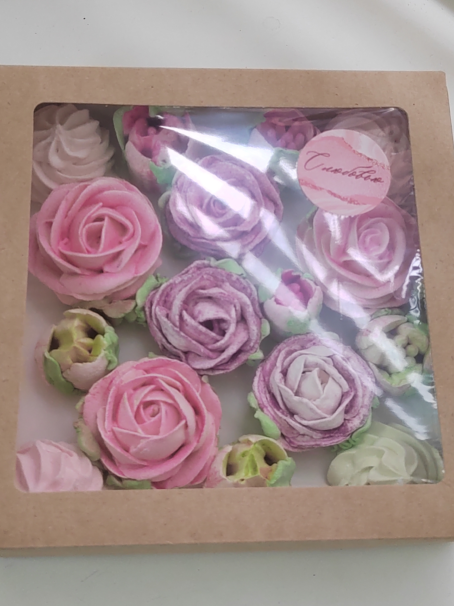 A close-up shows a square cardboard box, presumably a gift or bakery box, filled with decorative meringue cookies shaped like roses. The cookies are primarily in shades of pink and lavender, with some green accents presumably representing leaves. A clear plastic window in the box allows viewing of the contents. A circular sticker is on the upper-right section of the window. The contents of the box suggest a sweet confectionary product. This photo may have been taken in Votkinsk, Russia.
