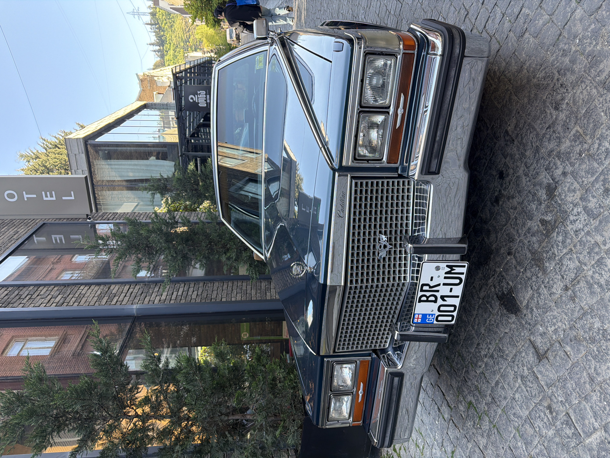 A blue Cadillac is parked on a cobblestone street in Tbilisi, Georgia. The license plate reads "BR-001-UM" with the Georgian flag. Behind the car is a modern building with a hotel sign. A couple of trees are visible. The sky is clear. A person walks by on the sidewalk.