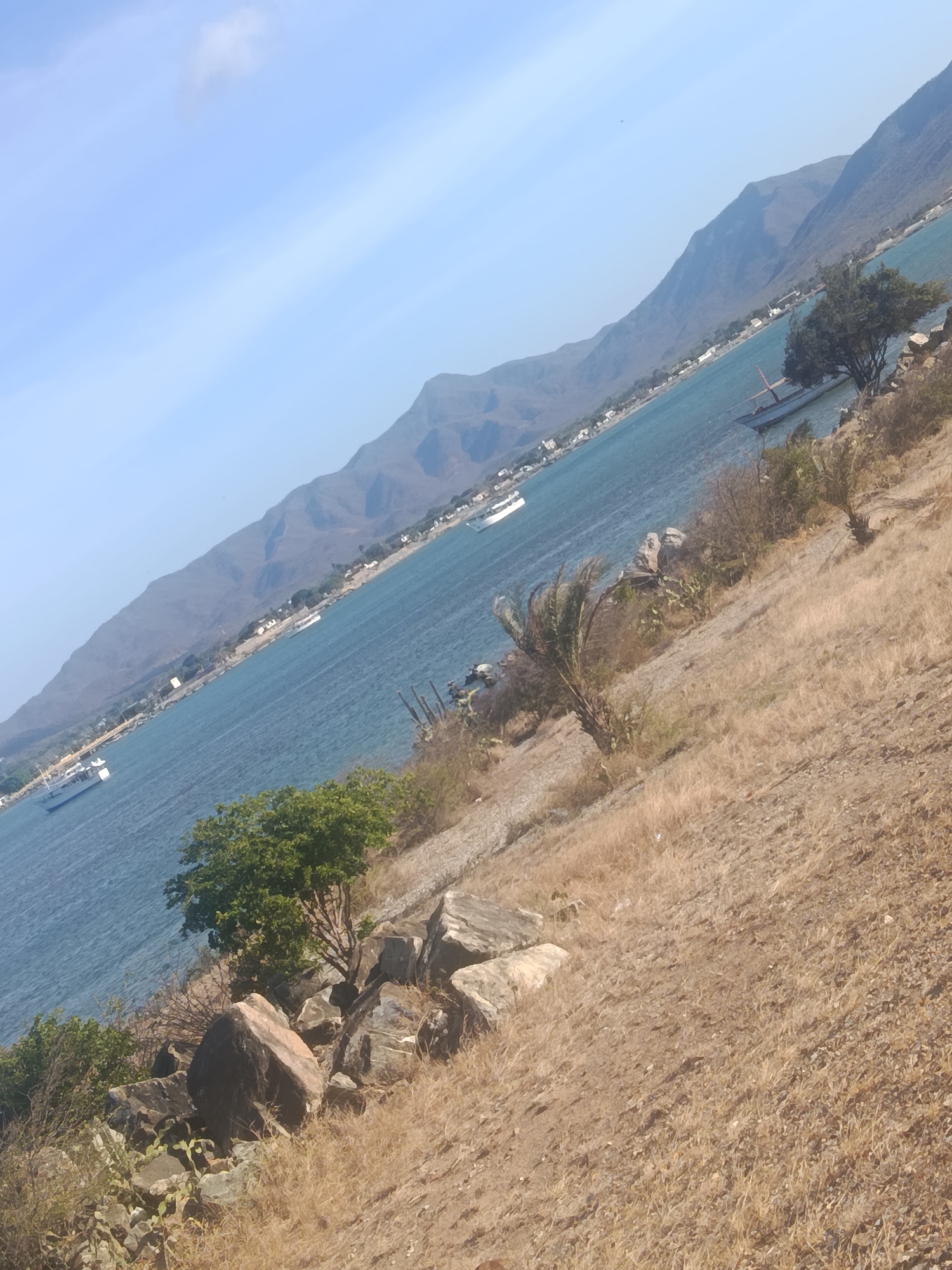The image depicts a coastal scene in Juan Griego, Venezuela. The foreground is dominated by a dry, grassy hillside with scattered rocks and vegetation, indicative of an arid environment. Moving towards the middle ground, the turquoise waters of the bay are visible, dotted with several boats. In the background, a mountain range stretches along the coastline, with buildings and settlements visible at the base of the mountains. The sky is clear blue with minimal cloud cover. The angle of the shot gives the impression of being taken from an elevated position on the hillside.
