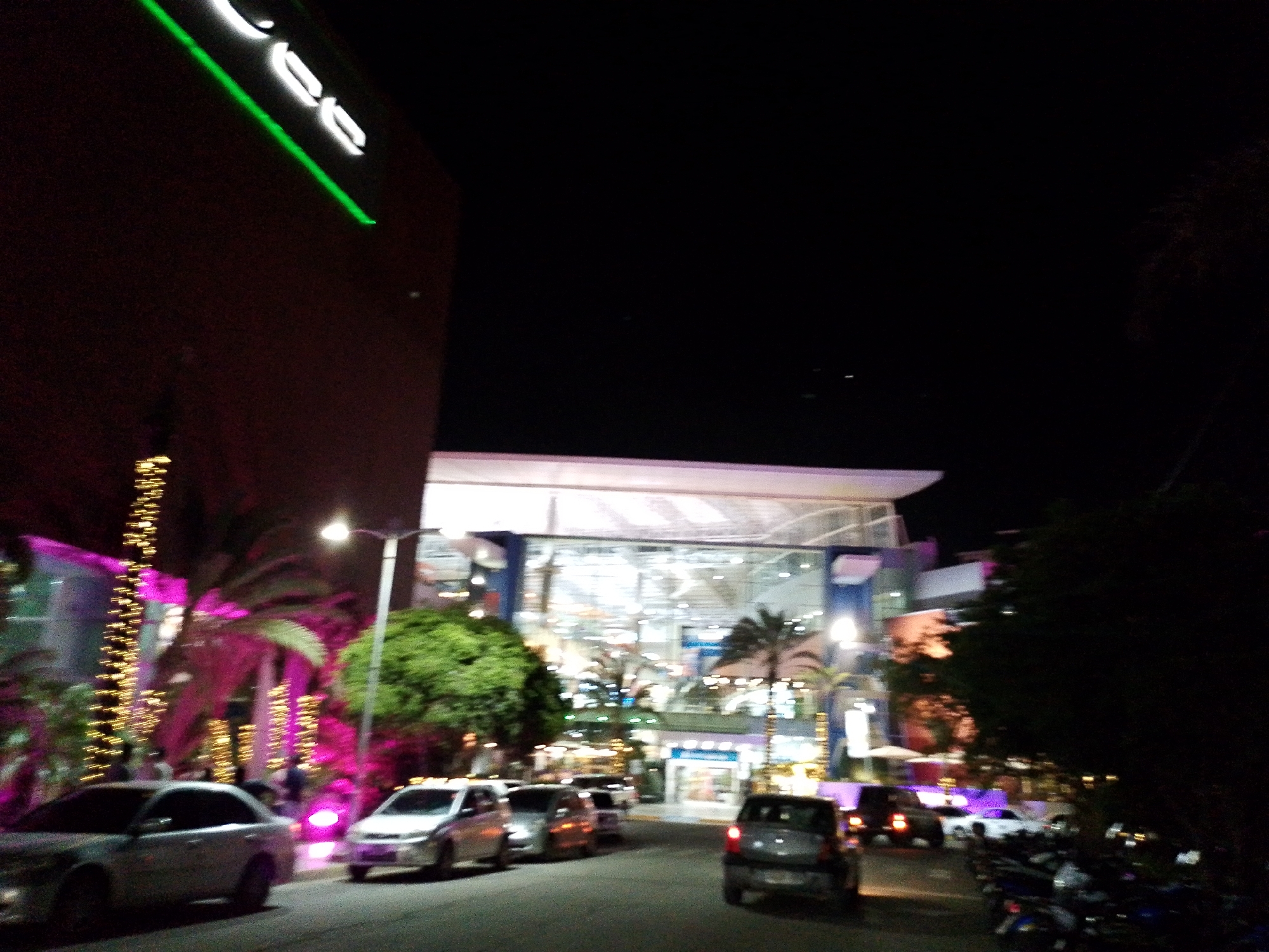 The image shows a street scene at night in Porlamar, Venezuela, likely near a shopping center. In the foreground, a silver sedan is parked on the right side of the street, facing away from the viewer. To its left, another silver sedan is visible, also parked. The street is paved and illuminated by streetlights and the lights from the buildings.
In the background, a modern building with a glass facade is illuminated, suggesting it is a commercial establishment, possibly a mall or a large store. Palm trees and decorative lighting, including strings of yellow and pink lights, are visible, indicating a festive or tropical setting. Several other vehicles, including cars and motorcycles, are parked or driving on the street. The overall atmosphere is that of a lively urban environment at night, with a focus on commercial activity and public spaces.