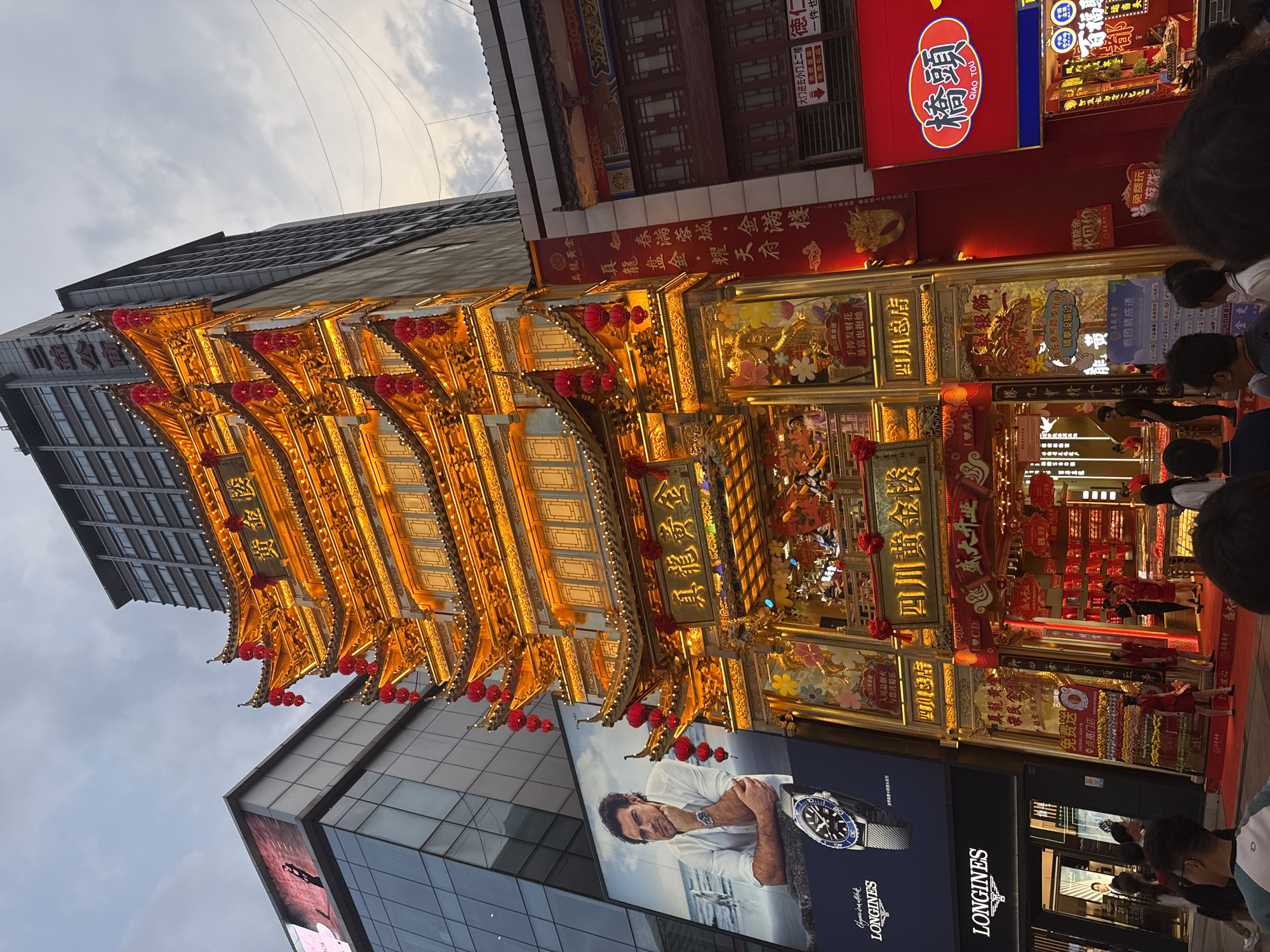 The image shows a street scene in Jinjiang District, China. A multi-tiered, brightly lit, gold-colored building with Chinese characters and red lanterns is prominent. A modern building with a large Longines advertisement featuring a man and a watch is visible below. People are walking on the street in front of the buildings. Multiple Chinese signs in red and gold are present.