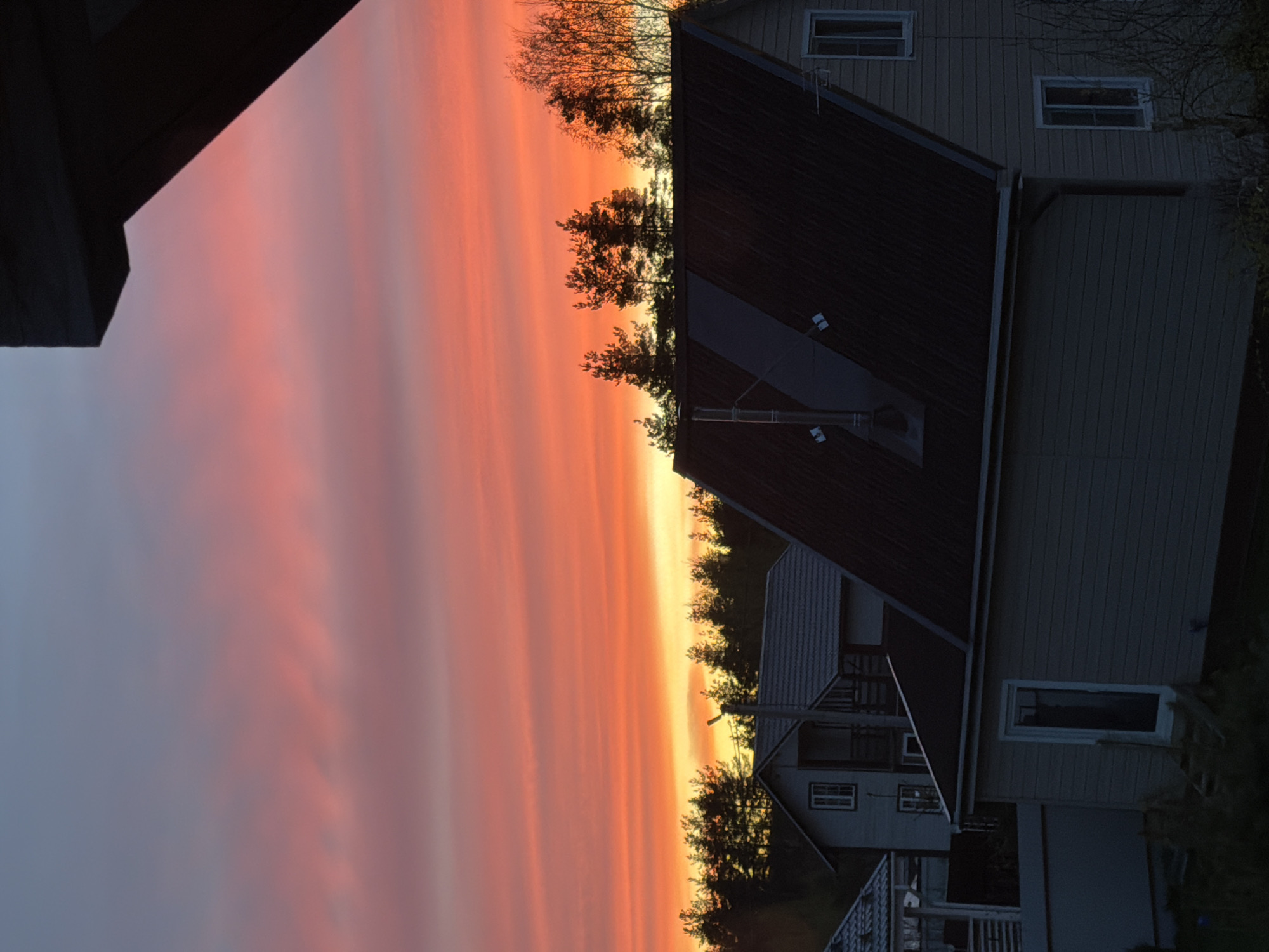 The image presents a sunset view in what appears to be a residential area. The sky exhibits vibrant hues of orange and pink, suggesting either sunrise or sunset. Silhouetted against the sky are trees and the rooftops of houses. The houses are of various sizes and designs, indicating a varied architectural style. One house has a dark roof and light-colored siding, and its windows are visible. A dark-colored overhang is present in the upper left corner of the image. The absence of people suggests a still moment captured during dawn or dusk. There are no identifying details to pinpoint the specific location in Steklyannyi, Russia.