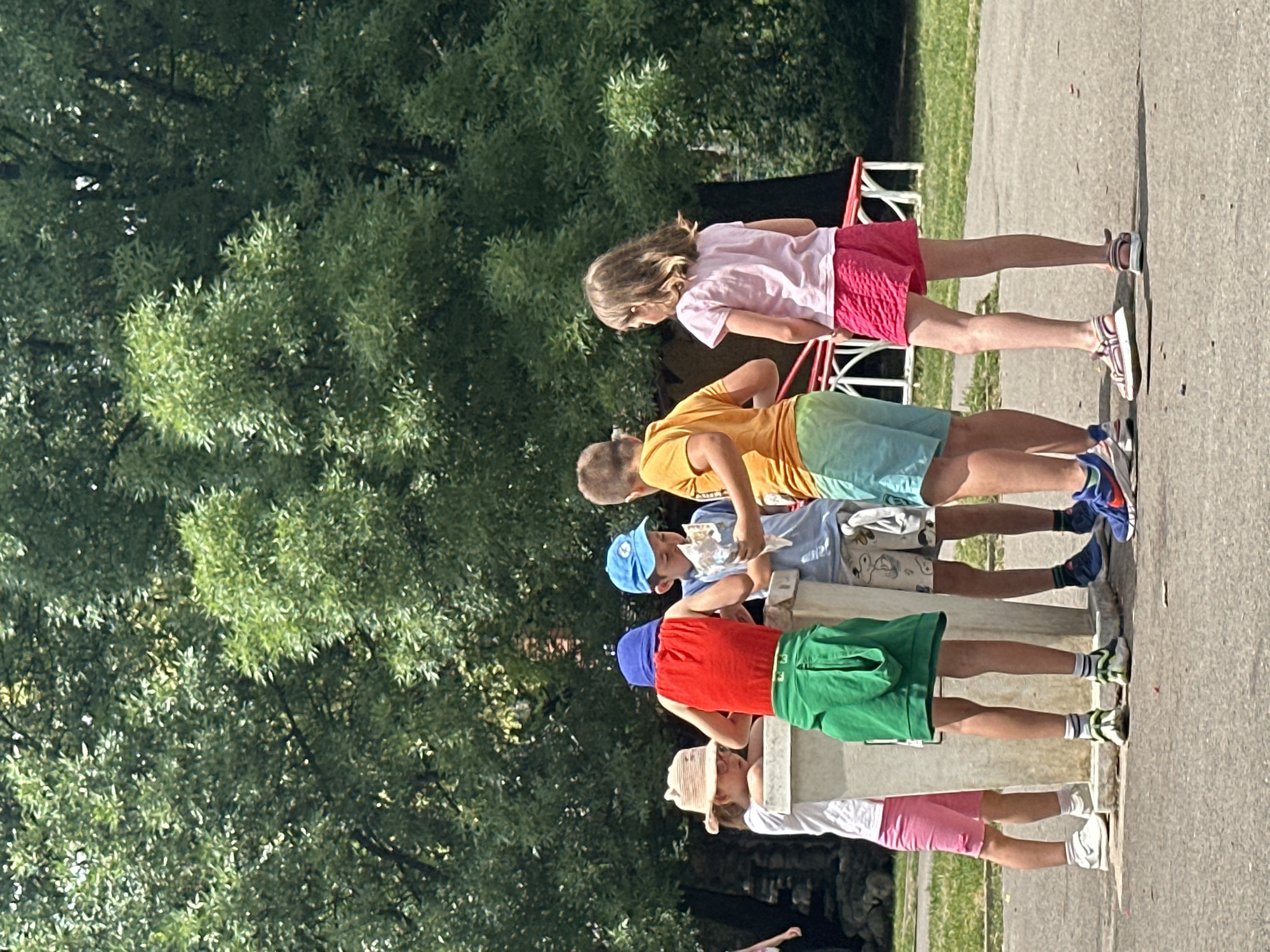 The image shows a group of children standing in a line outdoors. The children are dressed in casual summer clothing: t-shirts, shorts, and sneakers. One girl wears a pink t-shirt and red shorts. Next to her is a boy in an orange t-shirt and blue-green shorts. Following him is a boy in a blue t-shirt and patterned shorts, and another boy in a red t-shirt and green shorts. The last child visible is wearing a white t-shirt and pink shorts, with a straw hat. The children appear to be participating in some sort of outdoor activity or event, possibly a race or a game, as they are all facing the same direction and seem to be looking forward. The background consists of lush green trees and a paved surface, suggesting a park or an outdoor recreational area. The lighting indicates it is a sunny day, casting shadows on the ground. The children's expressions are not clearly visible, but the overall scene suggests a cheerful and active atmosphere. The location is stated to be Sofia, Bulgaria.