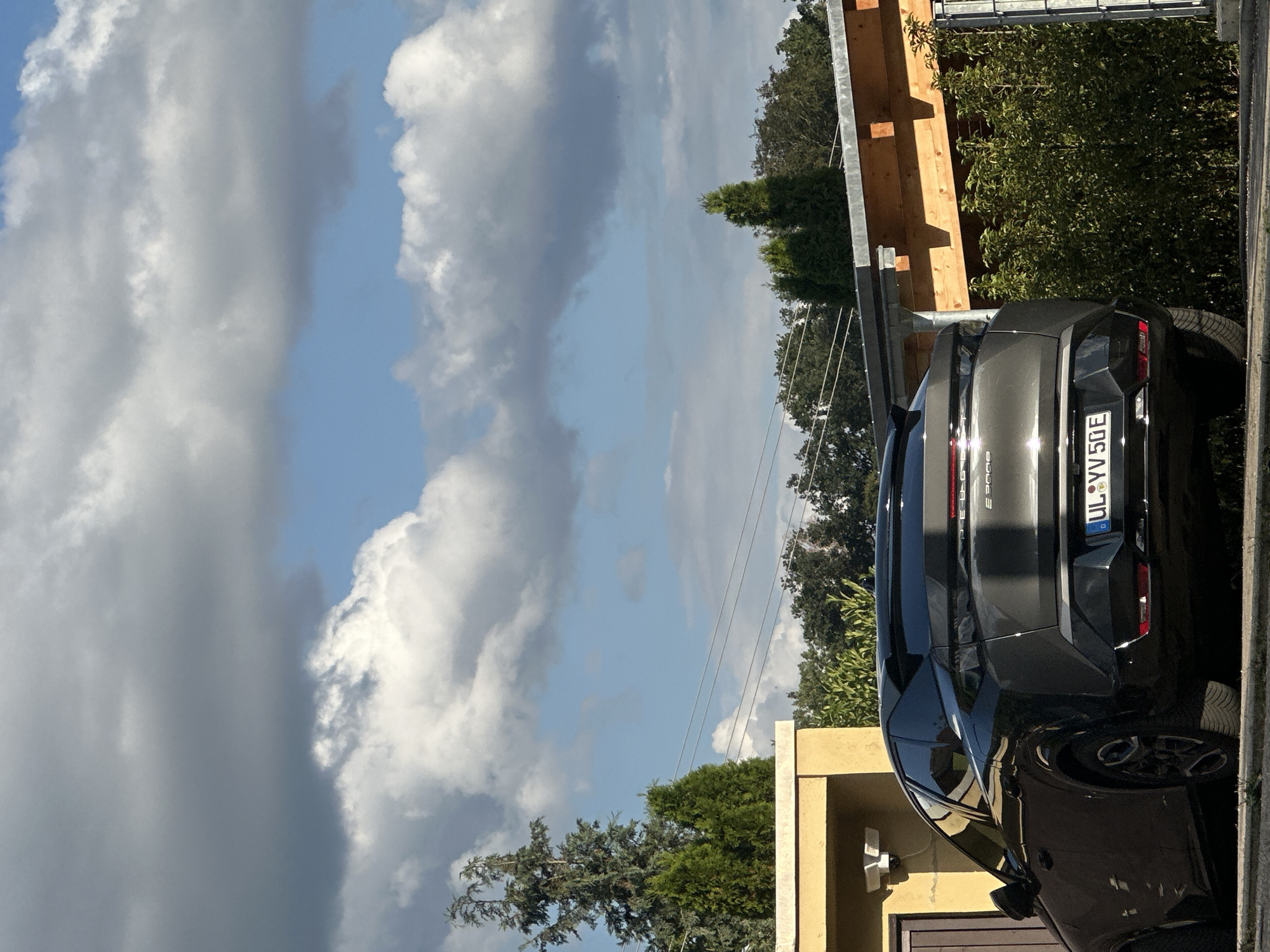The image shows the rear of a dark gray electric car parked outdoors, possibly in a driveway or parking lot. The car has a license plate that reads "UL VV 50E". In the background, there's a wooden structure, possibly a carport or pergola, with metal supports. Dense green trees and bushes are visible behind the structure. The sky is partly cloudy with a mix of dark and bright clouds against a blue sky, suggesting it might be late afternoon or early evening, with changing weather. The overall setting appears to be residential or a semi-rural area, with hints of greenery and natural elements. There are no people visible in the image. The scene is captured from a low angle, emphasizing the car and the sky. The lighting indicates it's daytime, with sunlight casting reflections on the car's surface.