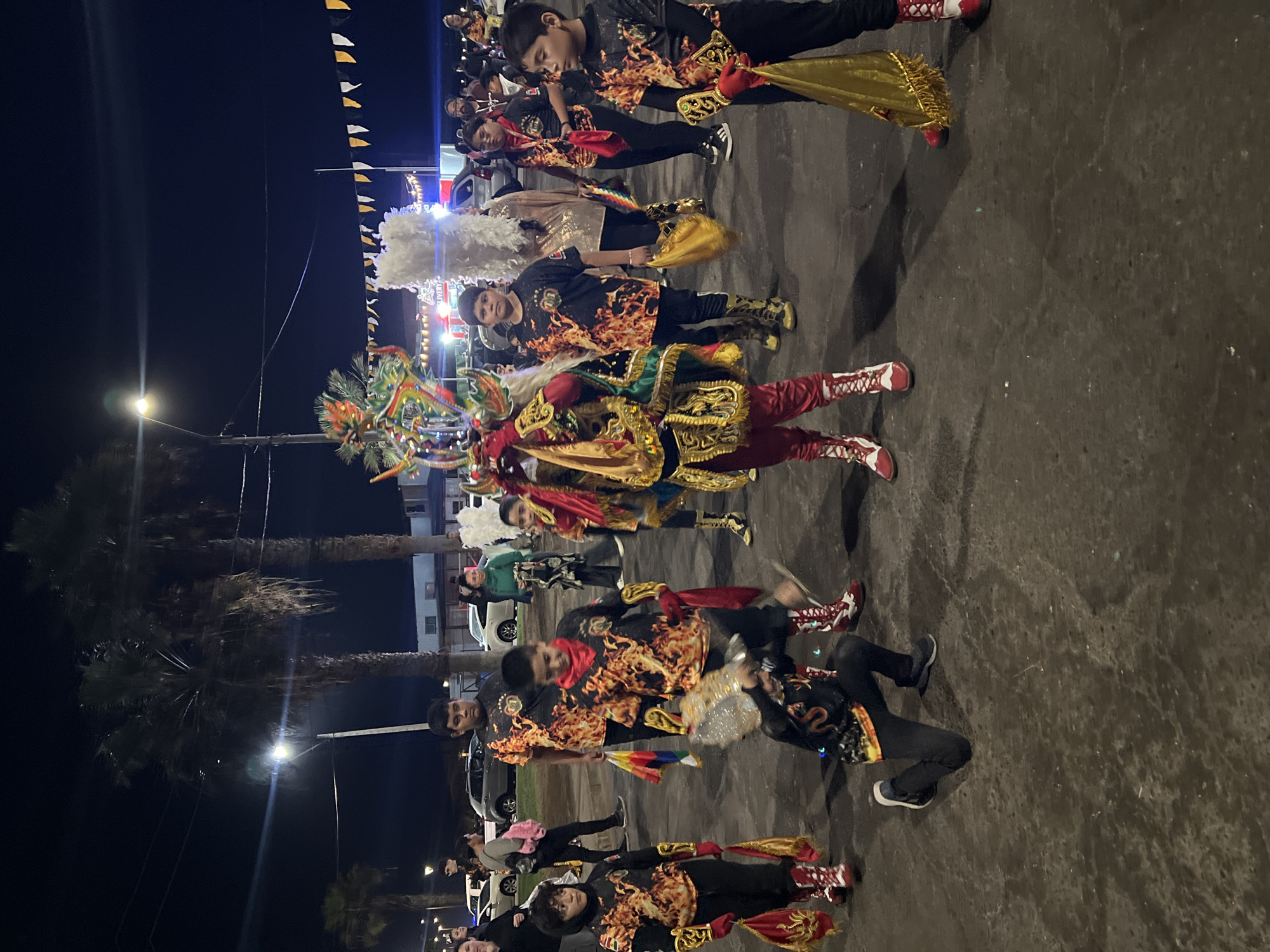 The image depicts a nighttime outdoor scene, likely a parade or cultural event in the city of Arica, Chile, as suggested by the palm trees characteristic of the region. In the foreground, several young individuals are dressed in vibrant costumes. One person in the center wears an elaborate headdress and a red and gold ornate costume, possibly a demon or mythical character. Other children are wearing black t-shirts with a flaming design, red and white boots, and some are holding golden bags or flags. One child is kneeling. In the background, there are more people, cars, buildings, and overhead streetlights, indicating an urban setting. String lights and pennants are visible in the distance, further suggesting a festive atmosphere. The ground appears to be asphalt or pavement, wet in some areas, reflecting the artificial lights.