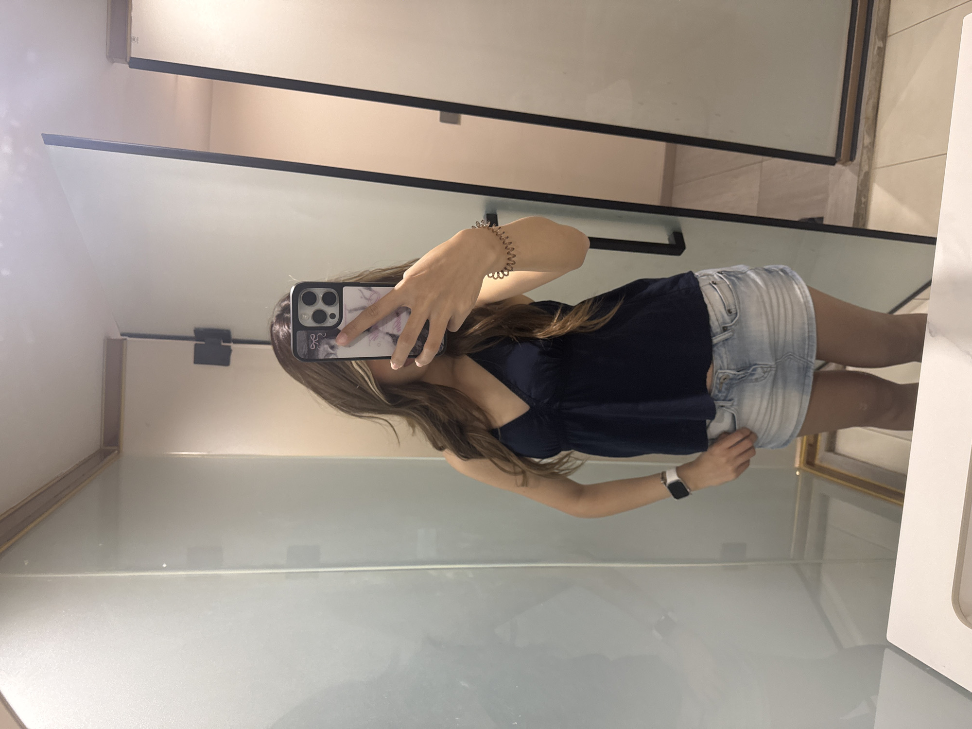 A woman is taking a selfie in what appears to be a public restroom or changing room. The woman is wearing a dark blue tank top and light blue denim shorts. She is holding a smartphone with a marble patterned case, obscuring her face. A bracelet adorns her left wrist and a smartwatch is on her right wrist. The room is enclosed by glossy white partition walls with black metal frames. The environment suggests an interior space, with potential elements of a commercial establishment present. There are no specific indications of Yuzhong District, China, within the image.