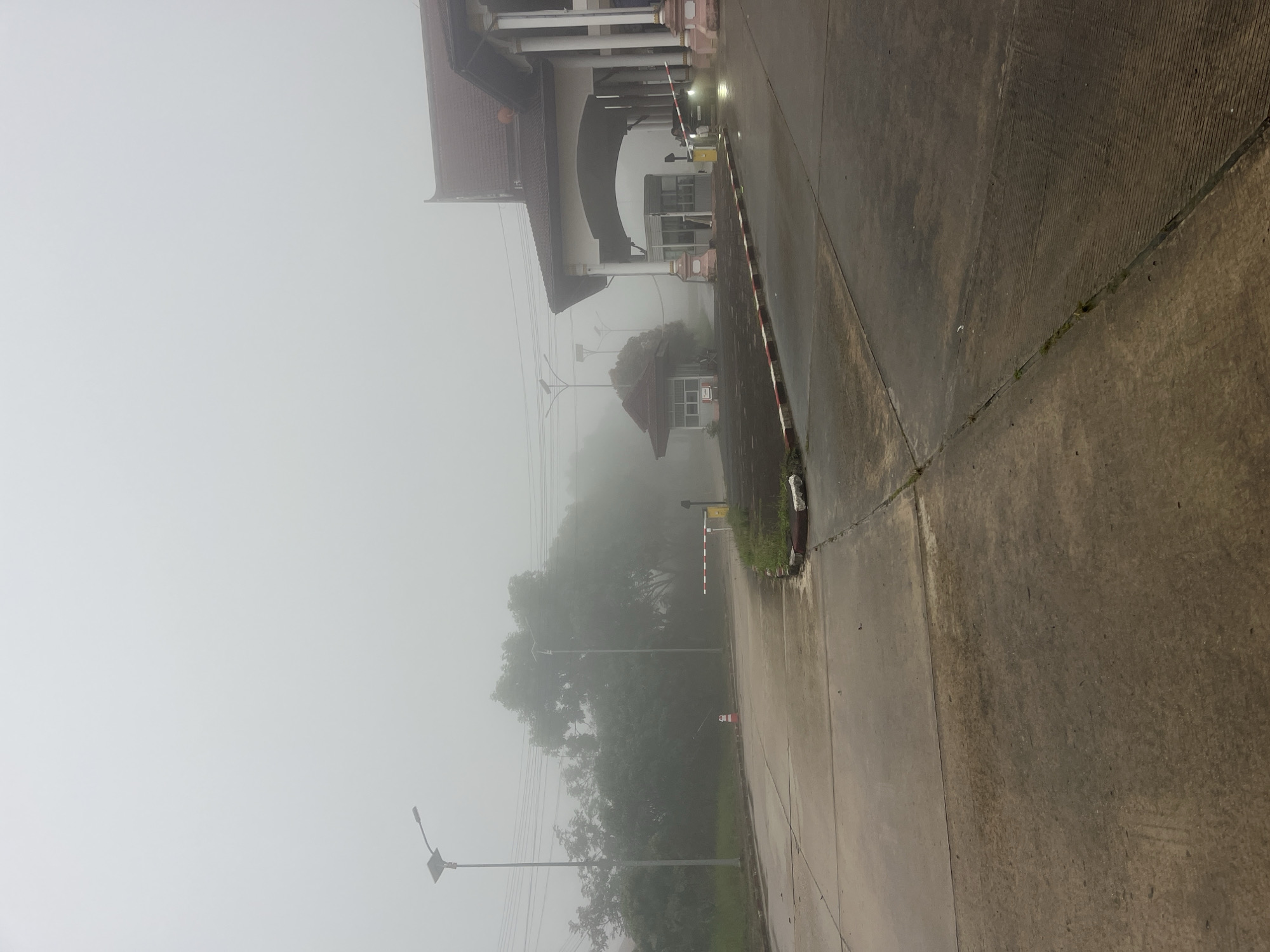 The image depicts a border crossing in the city of Xayden, Laos, shrouded in dense fog. The scene is dominated by a large, ornate building with a tiled roof, characteristic of the region's architecture, featuring grand columns and an arched entryway. In front of this building, a barrier gate is partially lowered, with a vehicle visible beyond it. To the left, a smaller, traditional-style building stands near the road. The road itself is paved and appears wet, possibly from recent rain or the damp fog. Lampposts with solar panels are strategically placed along the road, illuminating the area. Lush green trees are visible in the background, though their details are obscured by the thick mist. The overall atmosphere is quiet and somewhat isolated, with the fog creating a serene yet mysterious ambiance.