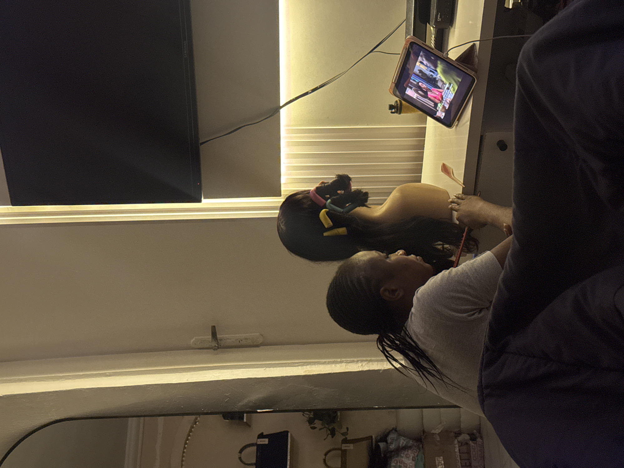The image shows a young woman with braided hair, wearing a grey t-shirt, sitting at a desk in a room. She is looking towards a tablet that is propped up on the desk, displaying a video with two women in it. Next to the tablet, there is a bottle and some black electronic equipment. To her left, a mannequin head with curlers in its hair is positioned, and the woman appears to be holding a pink comb or pencil. In the background, a large television screen is mounted on the wall, and to the far left, a tall mirror reflects a shelf with handbags and plants. The room is dimly lit by ambient lighting, with a strip of warm light above the desk and a brighter light source from the left. The scene suggests a domestic setting, possibly a bedroom or a personal study area, within a home in Abuja, Nigeria.