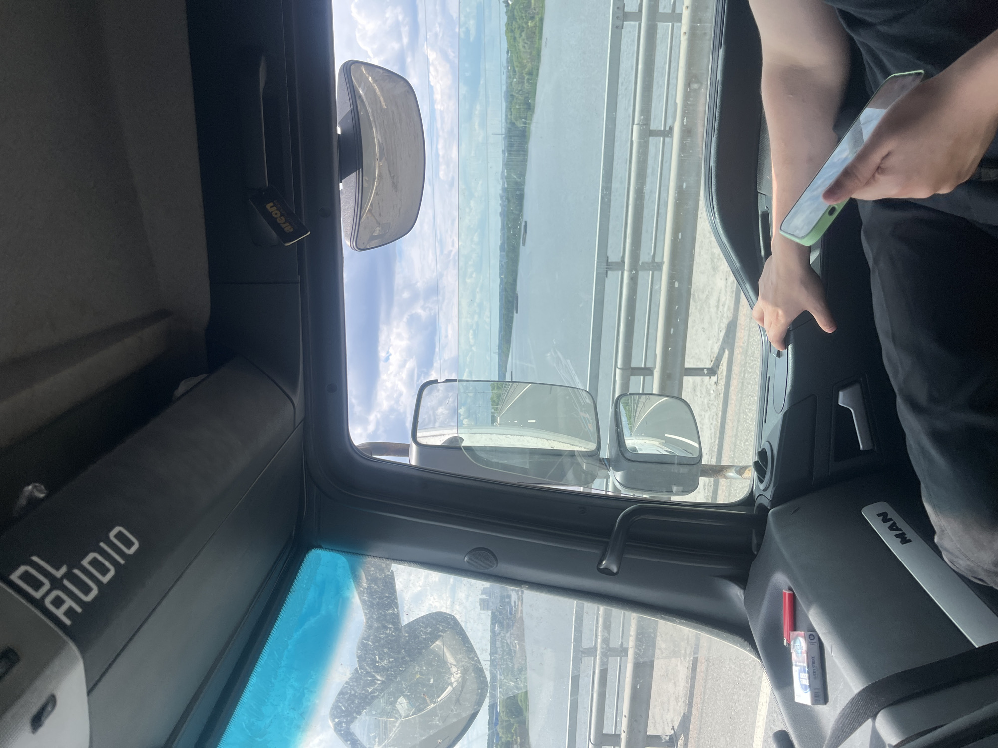 The image captures the interior of a truck cab, viewed from the passenger side looking forward and to the right. The truck is crossing a bridge over a wide body of water, likely a river, with land visible on the far bank. The sky is partly cloudy and bright.
Inside the cab, the dashboard is visible, with a "MAN" logo on the lower panel. On the dashboard, a pack of "Lucky Strike" cigarettes and a red lighter are placed on a textured surface. A person's hand is visible on the steering wheel on the right side of the frame, holding a light green smartphone. The person is wearing black clothing.
The truck's side mirror is positioned to reflect the road ahead and the surrounding environment. Mounted on the ceiling of the cab, above the windshield, is a black speaker or storage unit with white text that reads "DL AUDIO". A small air freshener, labeled "areon", is also attached to the ceiling. The windshield has a blue tint and appears to have some dirt or smudges. The overall impression is a glimpse into a moment of travel on a bridge, with everyday items present in the vehicle. The location is the bridge on the M-4 Don highway, near the village of Korobcheevo, Russia.