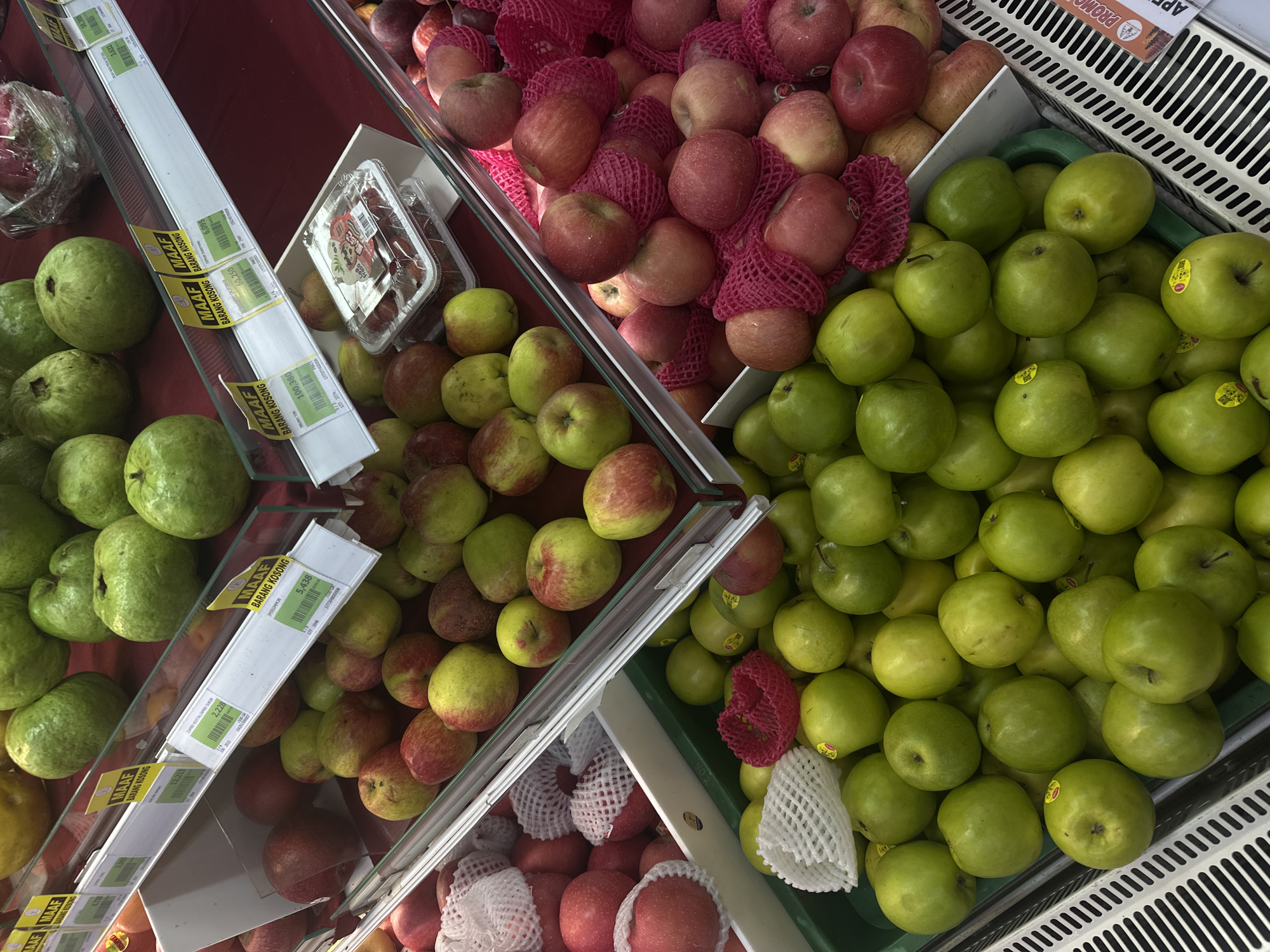 The image shows a produce display at a supermarket, likely in North Kuta, Indonesia. The display features a variety of fruits, predominantly apples and guavas, arranged on tiered shelves. The apples are in various shades of red and green, with some individually wrapped in pink mesh netting. The guavas are a vibrant green. Price tags and informational labels, some with the text "MAAF BARANG KOSONG," are visible on the shelves, indicating items that are out of stock. The overall environment appears to be a well-lit, commercial retail space, typical of a grocery store. There are no people visible in the image.