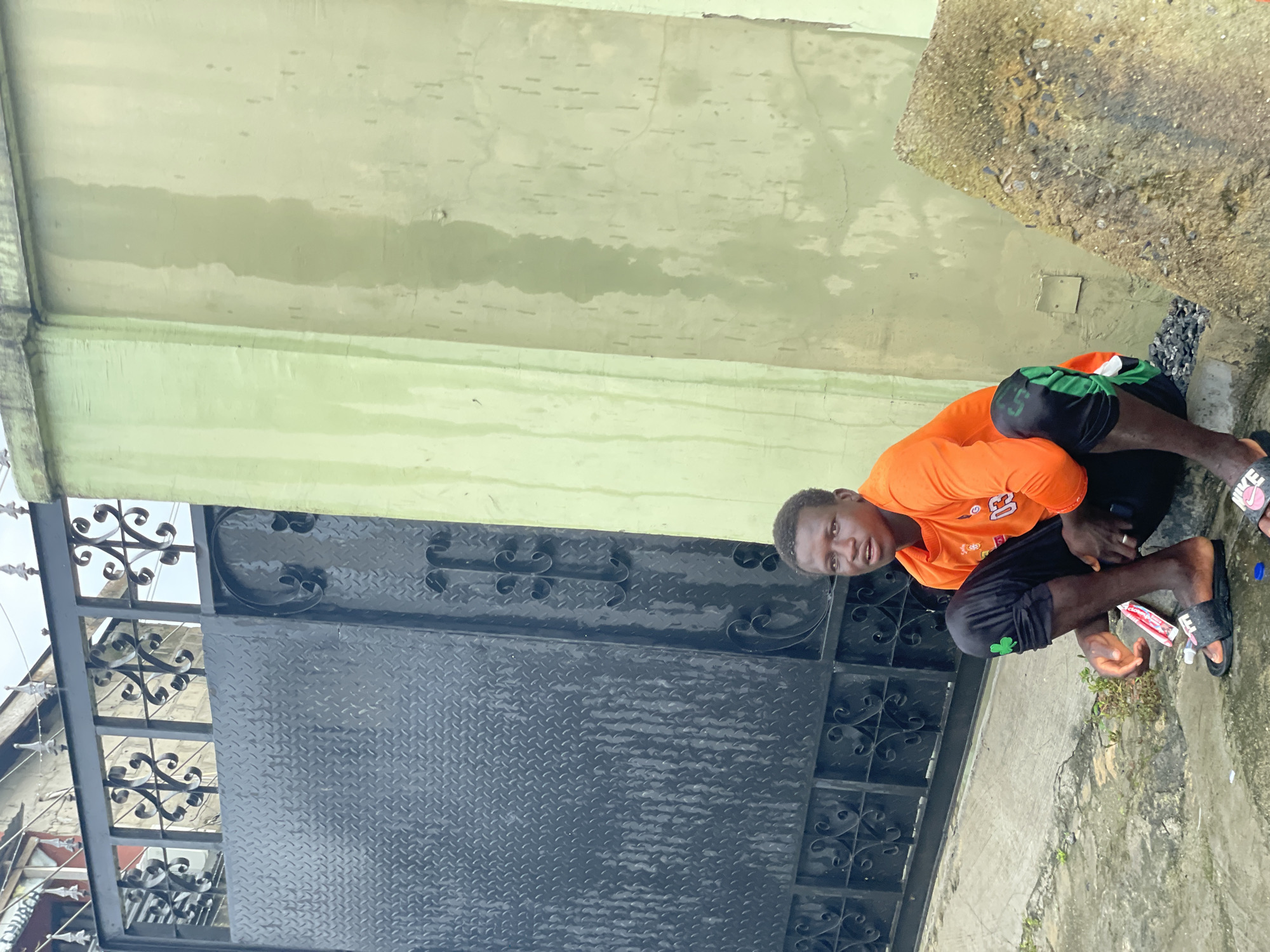 An outdoor scene in the city of Ado, Nigeria, depicts a young male crouching on a wet concrete surface in front of a building. The individual has dark skin, short dark hair, and is wearing an orange long-sleeved shirt with a partially visible number "03" and black athletic pants with green side stripes and a green shamrock emblem on the left thigh. He wears black Nike slide sandals and a silver ring on his left ring finger. His gaze is directed towards the viewer with a neutral expression, and his mouth is slightly open.
To the left of the individual is a dark grey metal gate. The lower section of the gate features a diamond plate pattern, while the upper section consists of ornate black scrollwork topped with spikes. The gate exhibits vertical streaks, consistent with water runoff or staining. Behind the gate, a light-colored building wall with a barred window is visible.
To the right of the individual is a light green painted concrete wall, which also displays significant vertical water stains and discoloration. Adjacent to the person's right, a large, rough-textured concrete slab rests on the ground. The ground beneath the individual is wet, uneven concrete, with small pieces of gravel near his left foot. A white and red wrapper is on the ground near his right hand, and a blue bottle cap is visible near his left foot. The overall environment suggests a humid or recently wet condition.