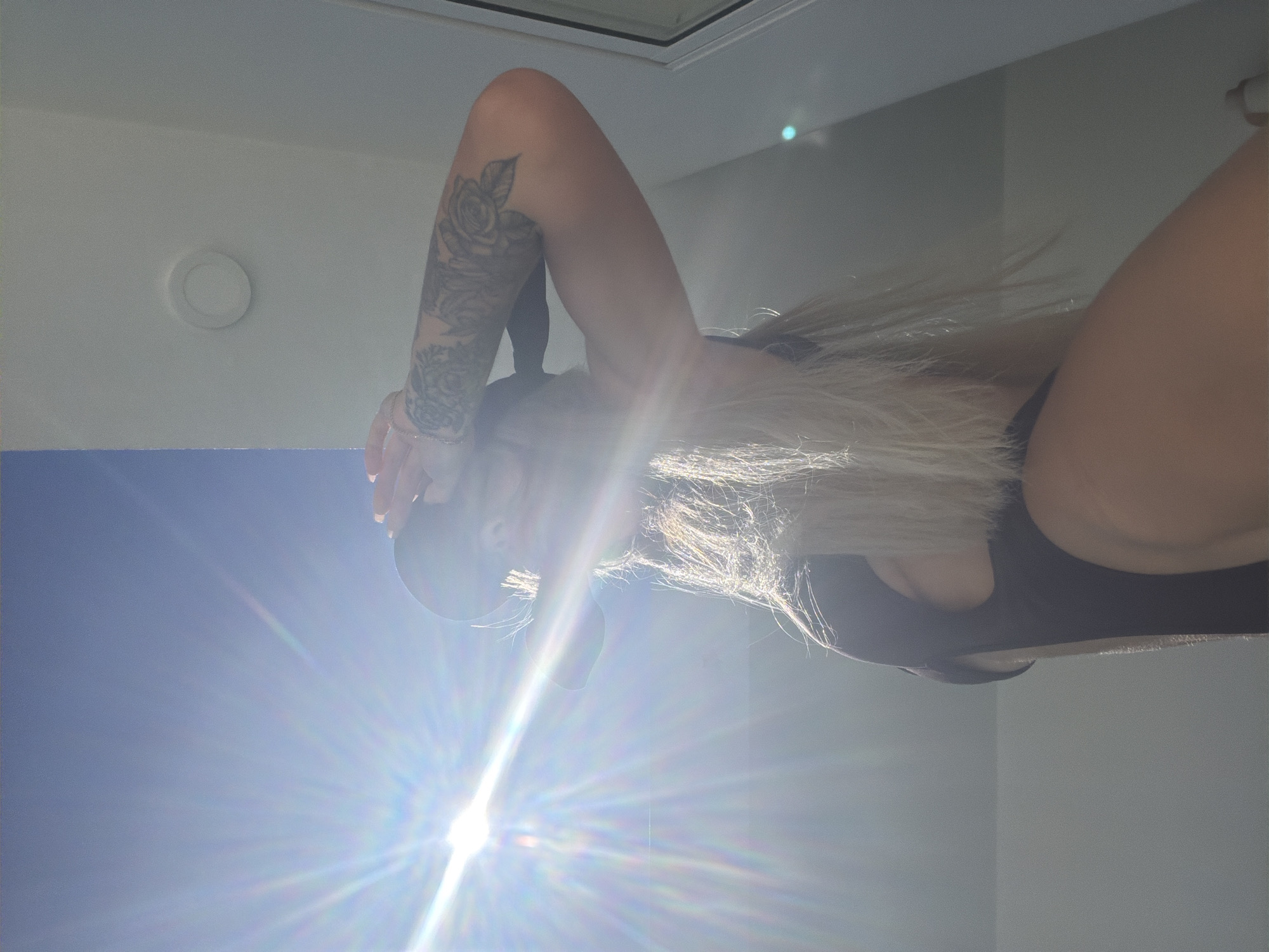 The image captures a low-angle, sun-drenched view of a woman wearing a black garment and a hat. She is standing outdoors, shielding her eyes from the bright sun with her hand. Her arm is adorned with tattoos. The environment appears to be a bright, sunny day with a clear blue sky. There is a white ceiling-like structure visible above and to the right of the woman, with a circular light fixture embedded in it. The sunlight creates strong lens flares and highlights, obscuring some details of the scene. The location is specified as Baan Bang Rak, Thailand.