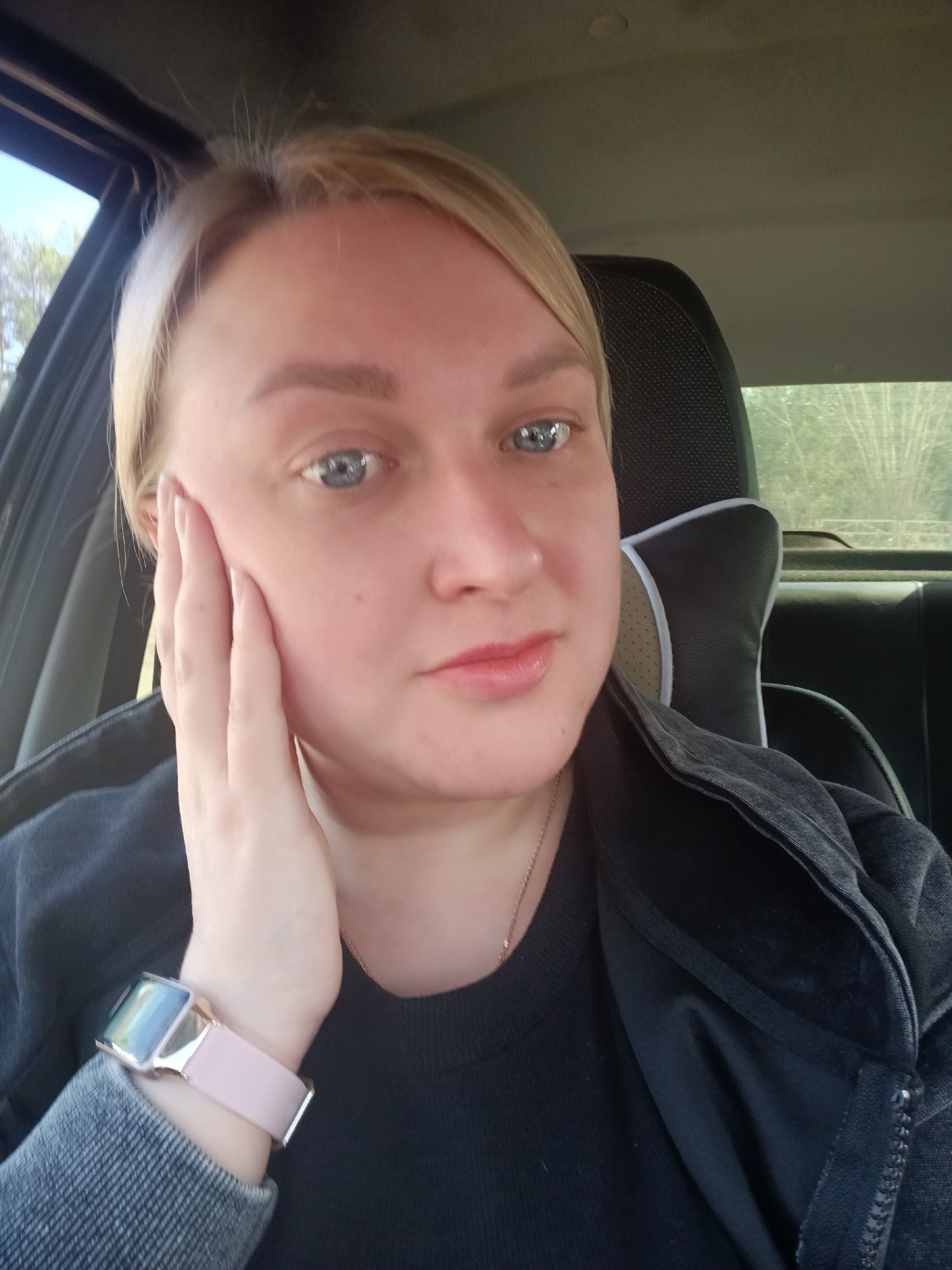 A fair-skinned woman with blonde hair and blue eyes is captured in a close-up, likely a selfie, inside a car in Chaikovsky, Russia. She is looking directly at the camera with a gentle, calm expression and a subtle smile. Her right hand rests lightly on her right cheek.
She is dressed in a black long-sleeved top, possibly a t-shirt or sweater, and an open dark grey or black jacket with visible stitching and a zipper detail on the front. A thin, delicate gold-colored necklace is visible around her neck. On her left wrist, she wears a smartwatch featuring a rectangular metallic gold-colored frame and a light pink band.
The background reveals the interior of the car, including a black seat headrest with a patterned texture and light-colored trim, and a light grey, textured ceiling. Through the car windows, blurred green foliage and glimpses of the sky are visible, indicating that the photo was taken during daylight hours. The natural light softly illuminates her face. There is no visible text in the image.